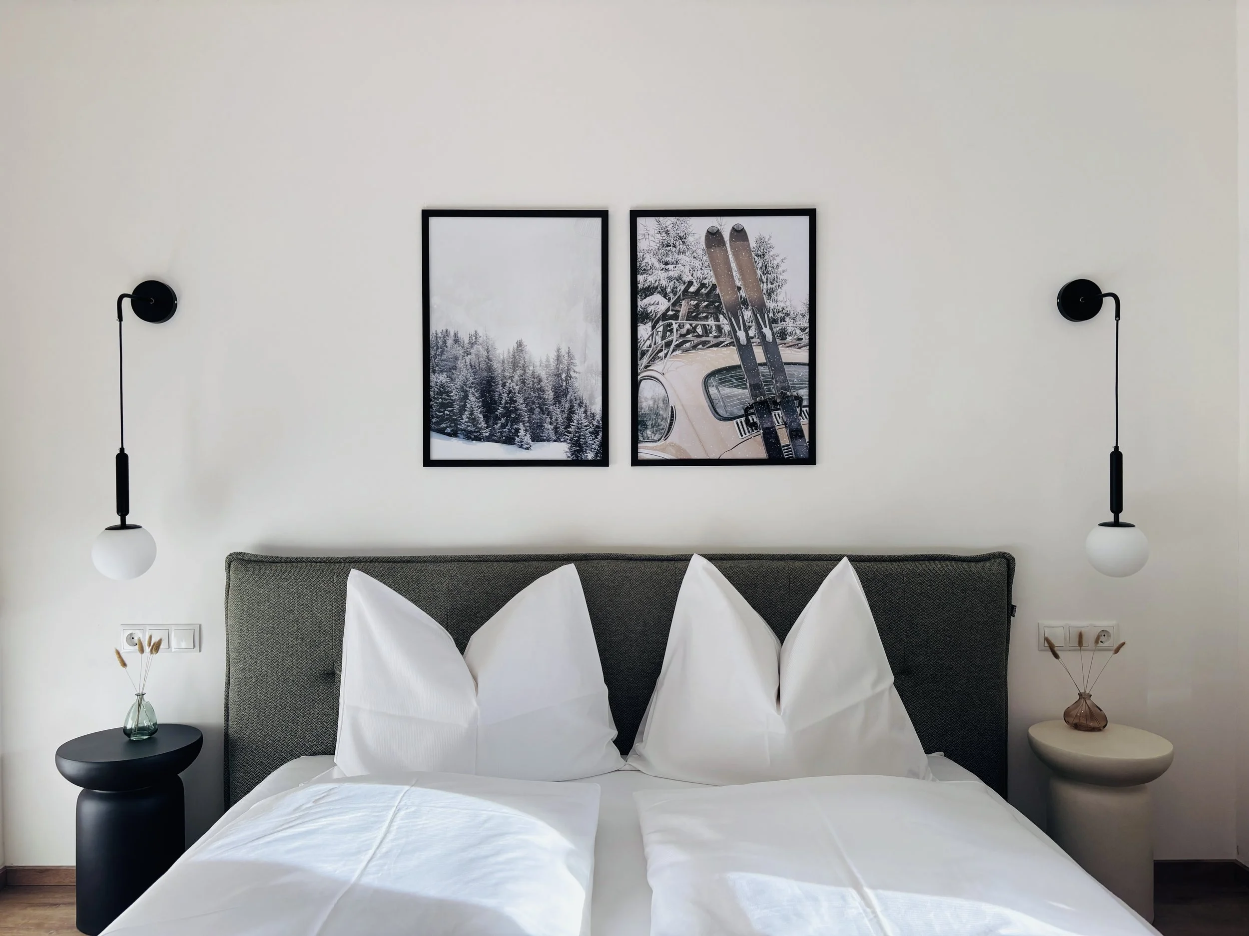 Schlafzimmer mit zwei weißen Betten, grauem Kopfteil, Wandlampen, zwei Bilder mit Winter- und Skithema, kleine Nachttische mit Dekogläsern
