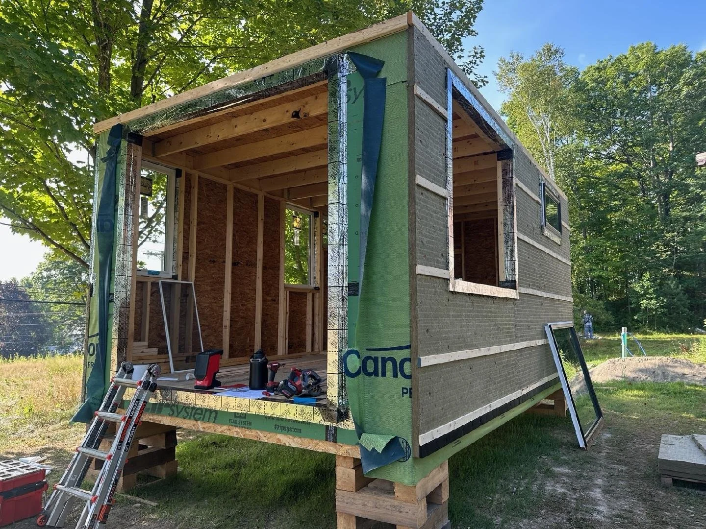 Small house under construction with wooden framing, insulation, and windows installed, surrounded by trees and construction tools outside.