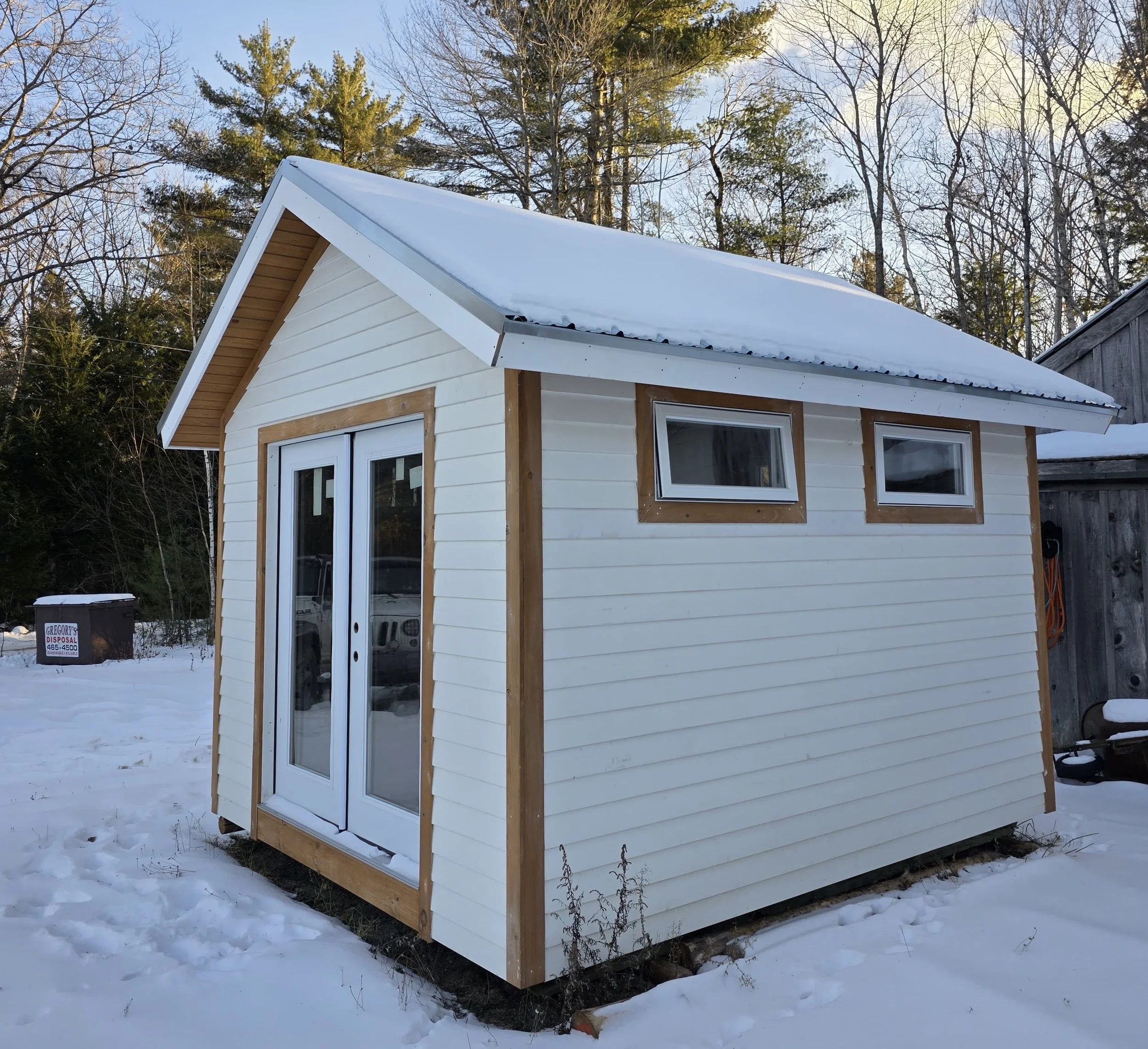 Small white shed with double doors, two small horizontal windows, in a snowy backyard with trees and a fence.