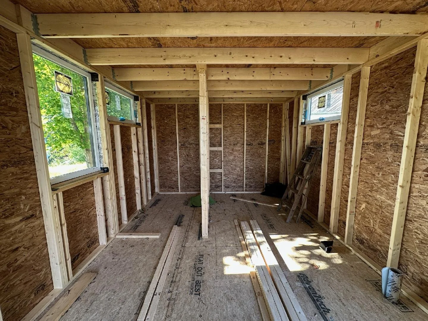 Interior of a room under construction with exposed wooden framing, OSB walls, and windows on two sides, sunlight coming through, construction materials and a ladder inside.