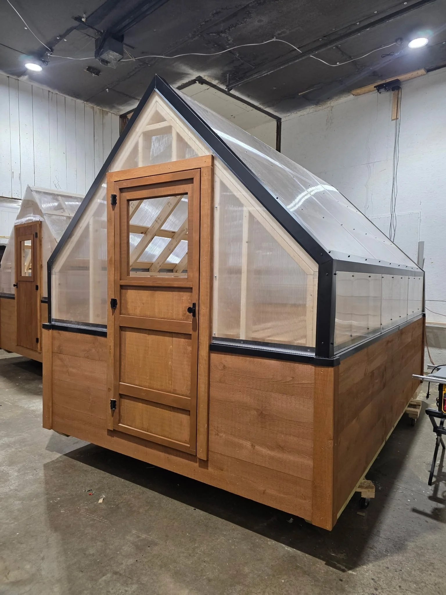 A small greenhouse with a wooden frame, transparent plastic panels, and a wooden door, situated indoors.