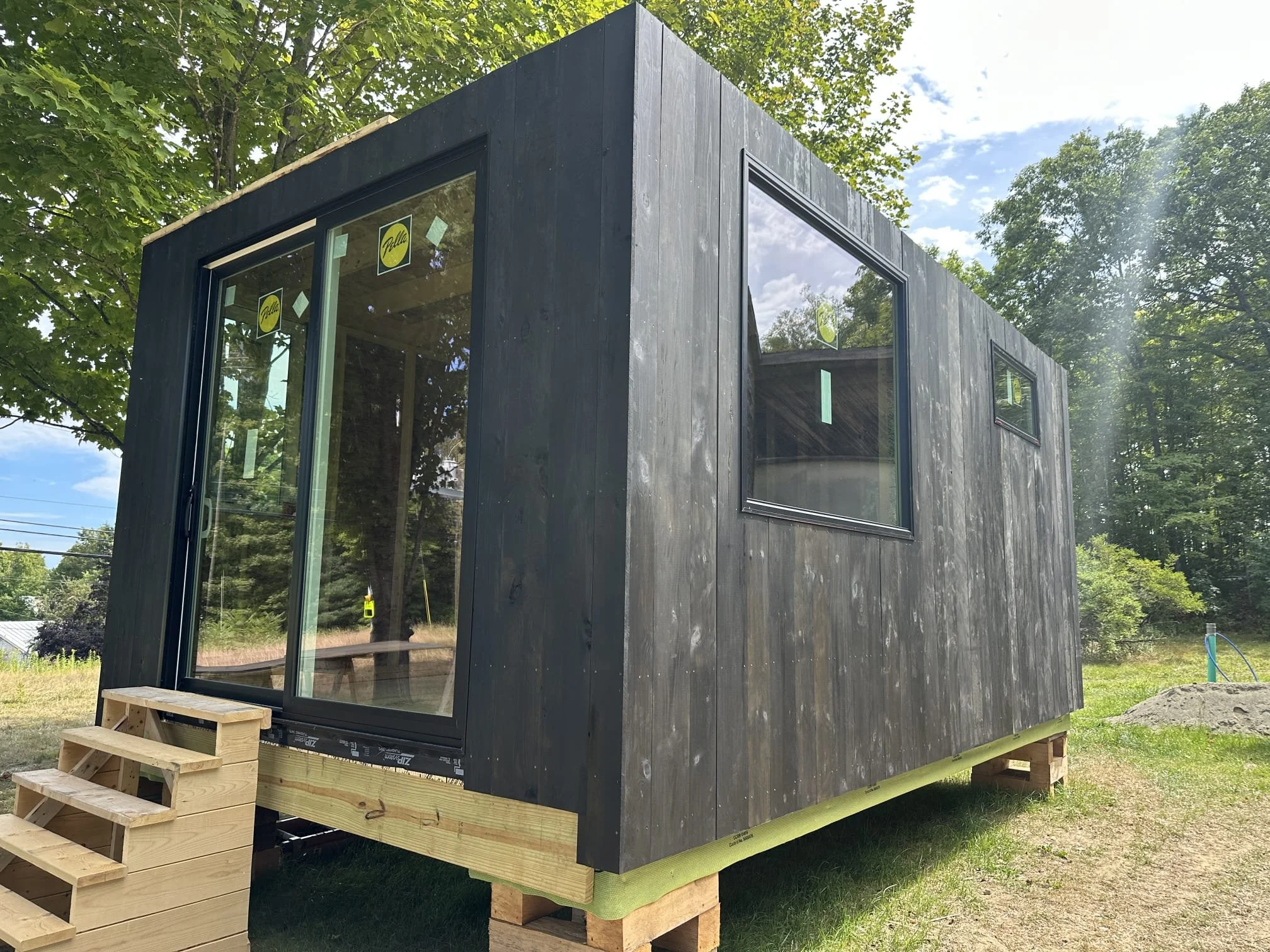 Small black wooden house or tiny home on a raised foundation with stairs outside, surrounded by trees and a grassy area, under a partly cloudy sky.