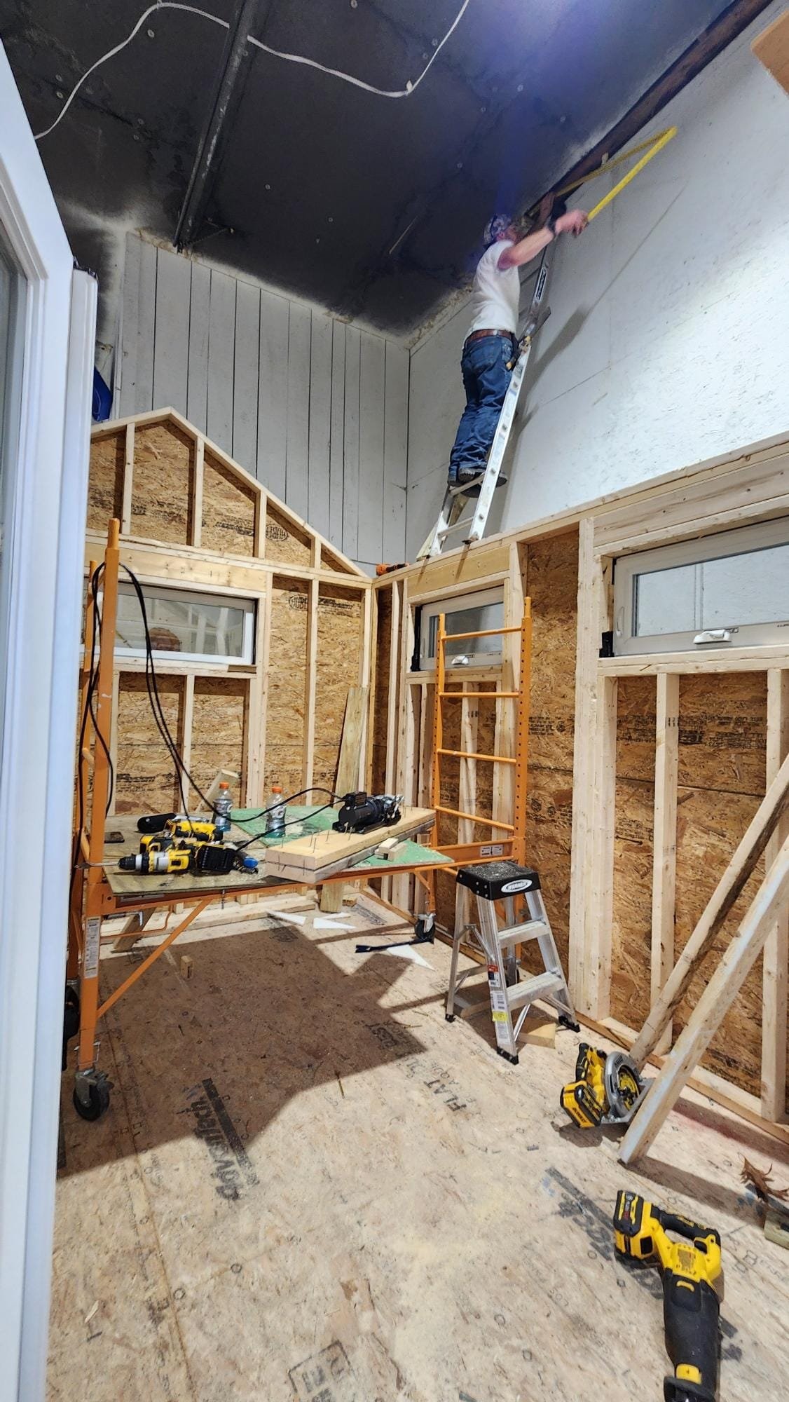 A person working on a home renovation, using a ladder to reach the ceiling, with construction materials and tools such as drills, a saw, and a stepladder in the room.