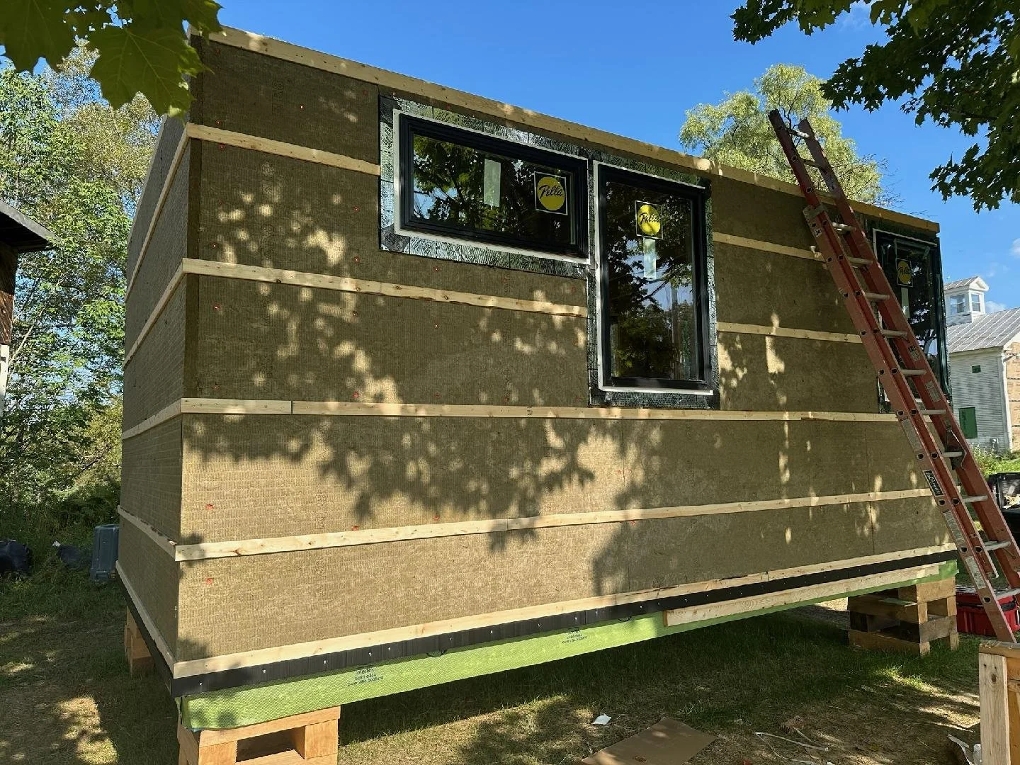 A small house under construction with beige insulation panels on its exterior, black-framed windows, a ladder leaning against the wall, and green weatherproofing on the bottom edge, all set outdoors on a sunny day with trees around.