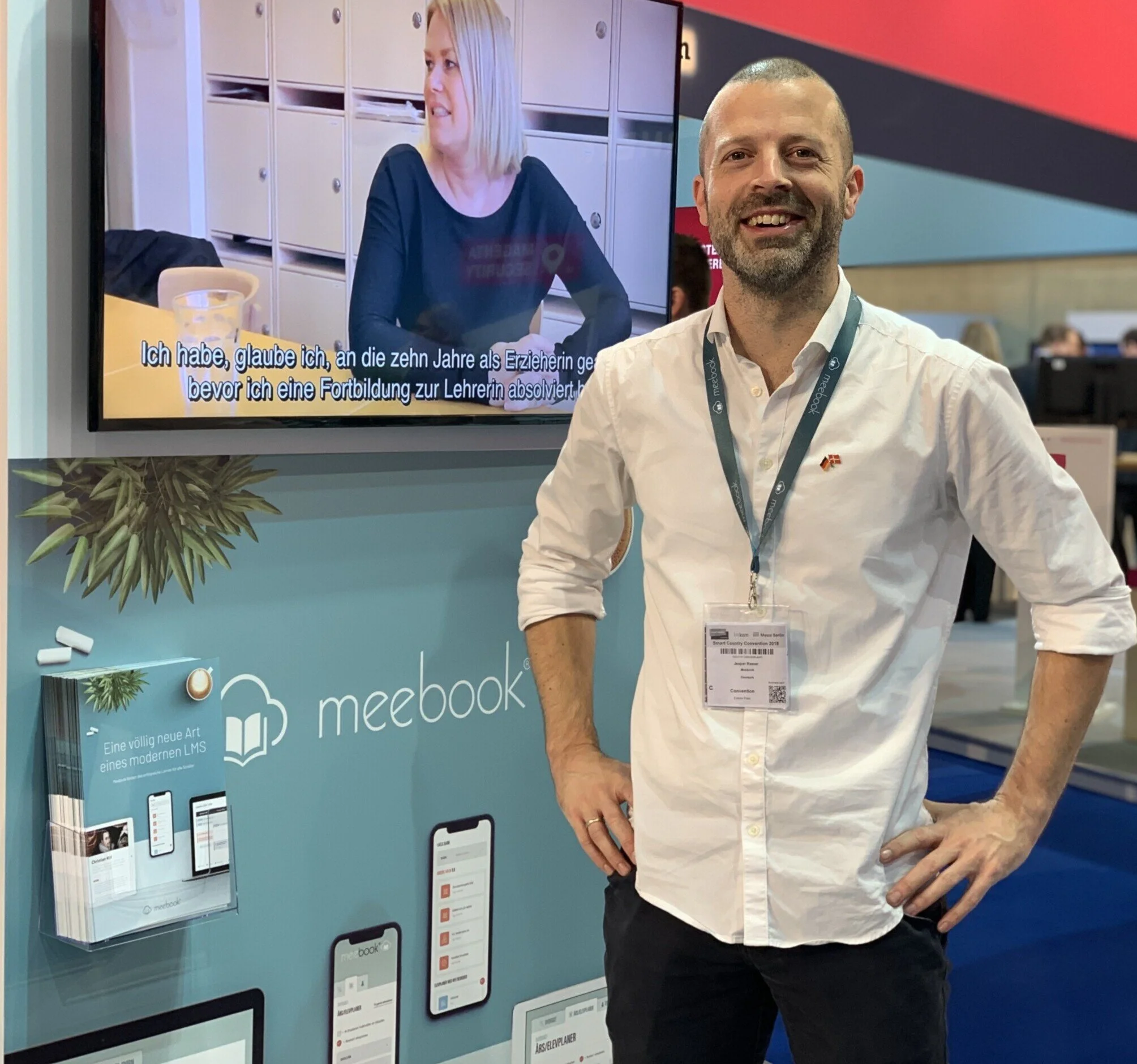 Ein Mann mit weißen Hemd und Badge posiert auf einer Messe neben einem Stand für meebook. Im Hintergrund ein Bildschirm mit einer Frau, die sich in einem Gespräch befindet, inklusive deutsche Untertitel.