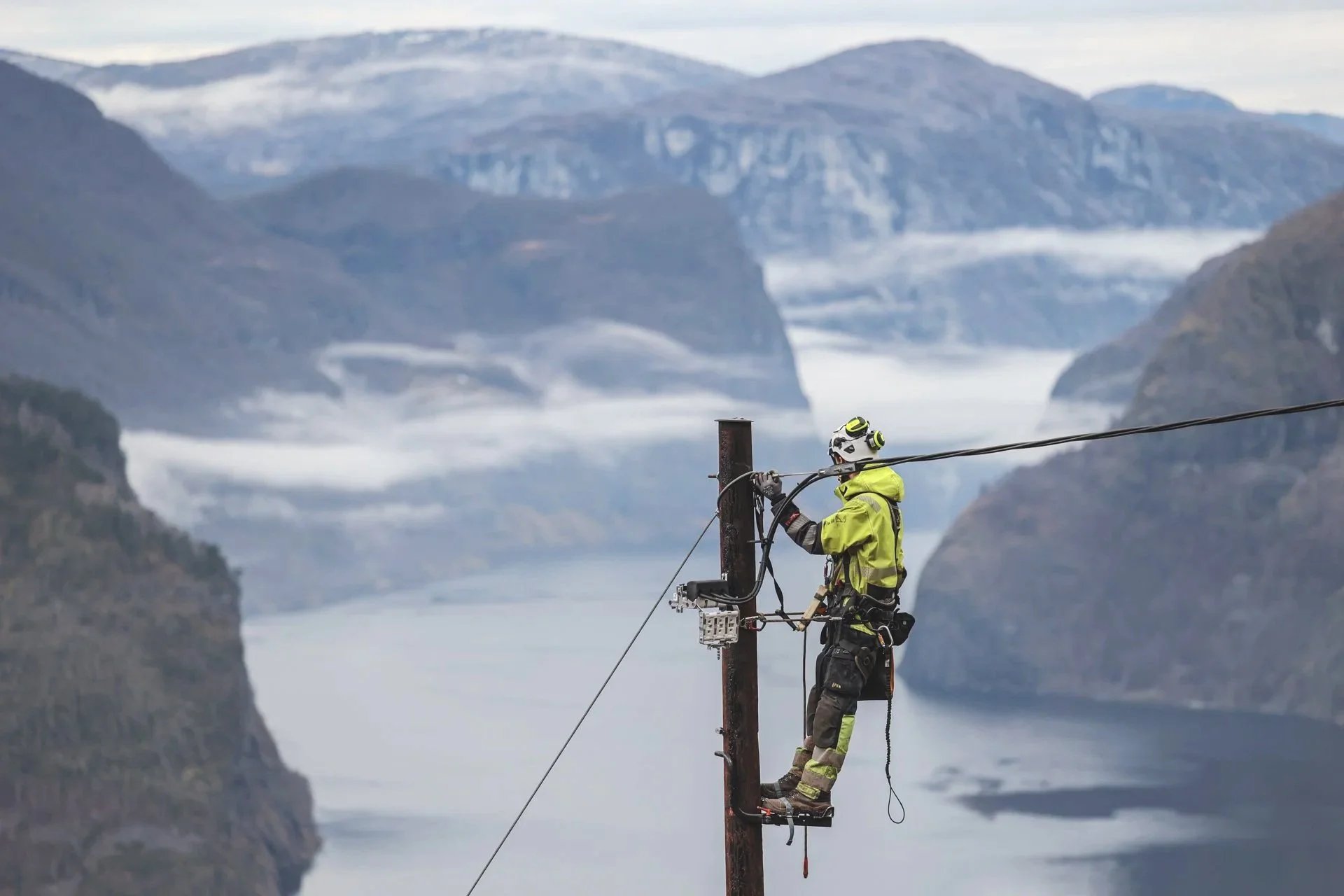 En person i arbeid på en kraftledning står på en tpop som er festet til en pole, med en fjord og fjell i bakgrunnen.