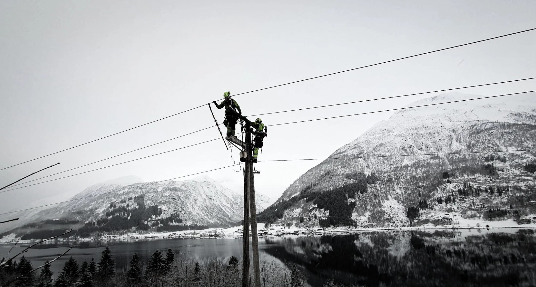 To nøkkelarbeidere jobber på en strømledning med snødekte fjell i bakgrunnen.
