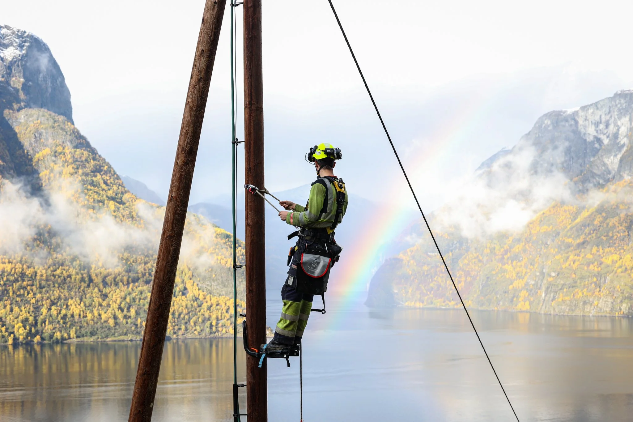 En mann iført verneutstyr som står i en stolpe, med en vakker fjord og fjell i bakgrunnen, der en regnbue er synlig.