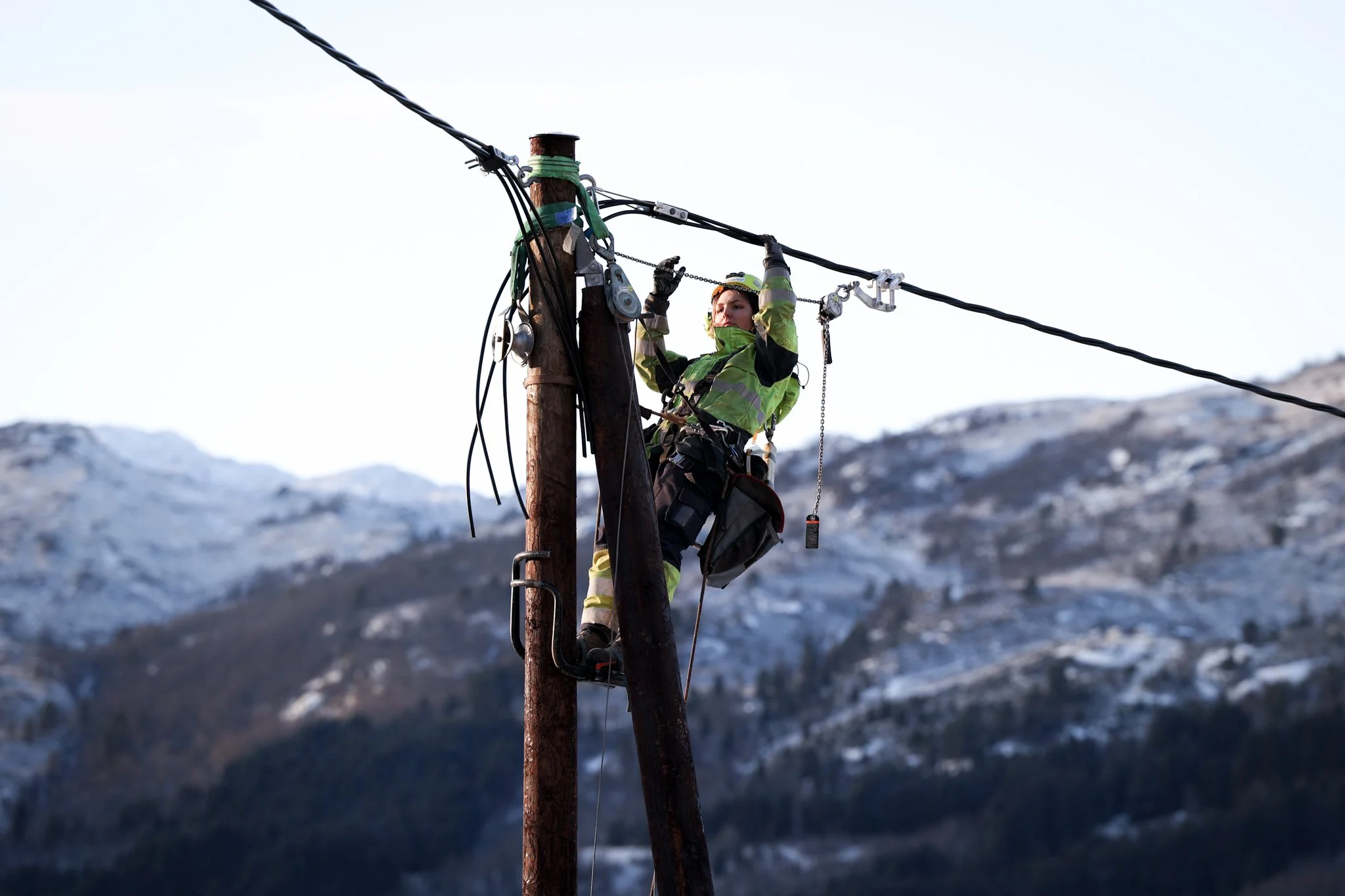En person i arbeidstøy står oppå en stolpe og reparerer en elektrisk ledning med fjell i bakgrunnen.