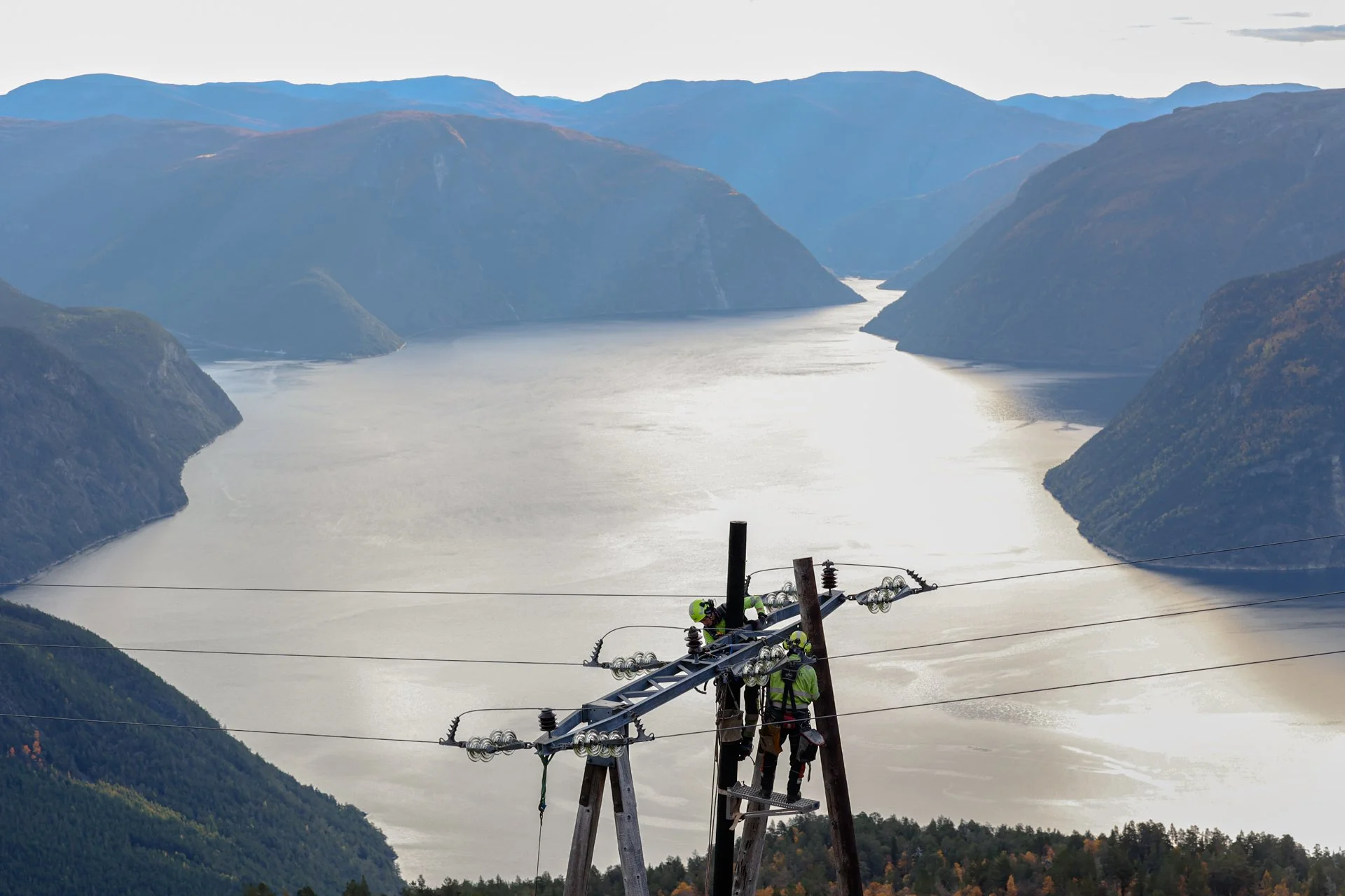 Elektrikarbeidere som arbeider på en kraftlinje med fjellandskap og innsjø i bakgrunnen.
