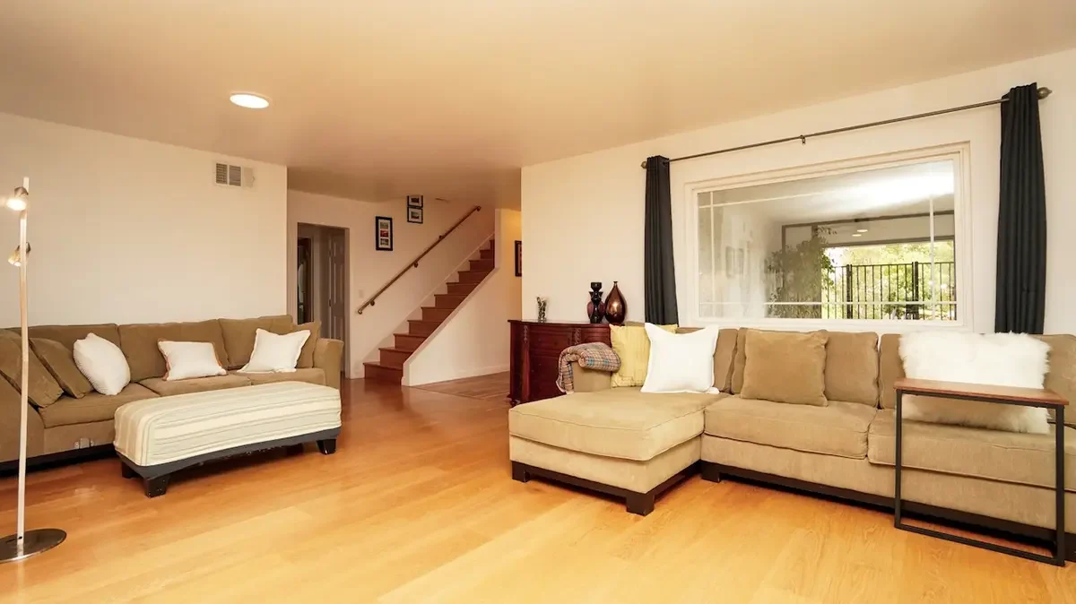 Living room with two beige sofas, wooden flooring, a small side table, curtains, and a window with a porch view.