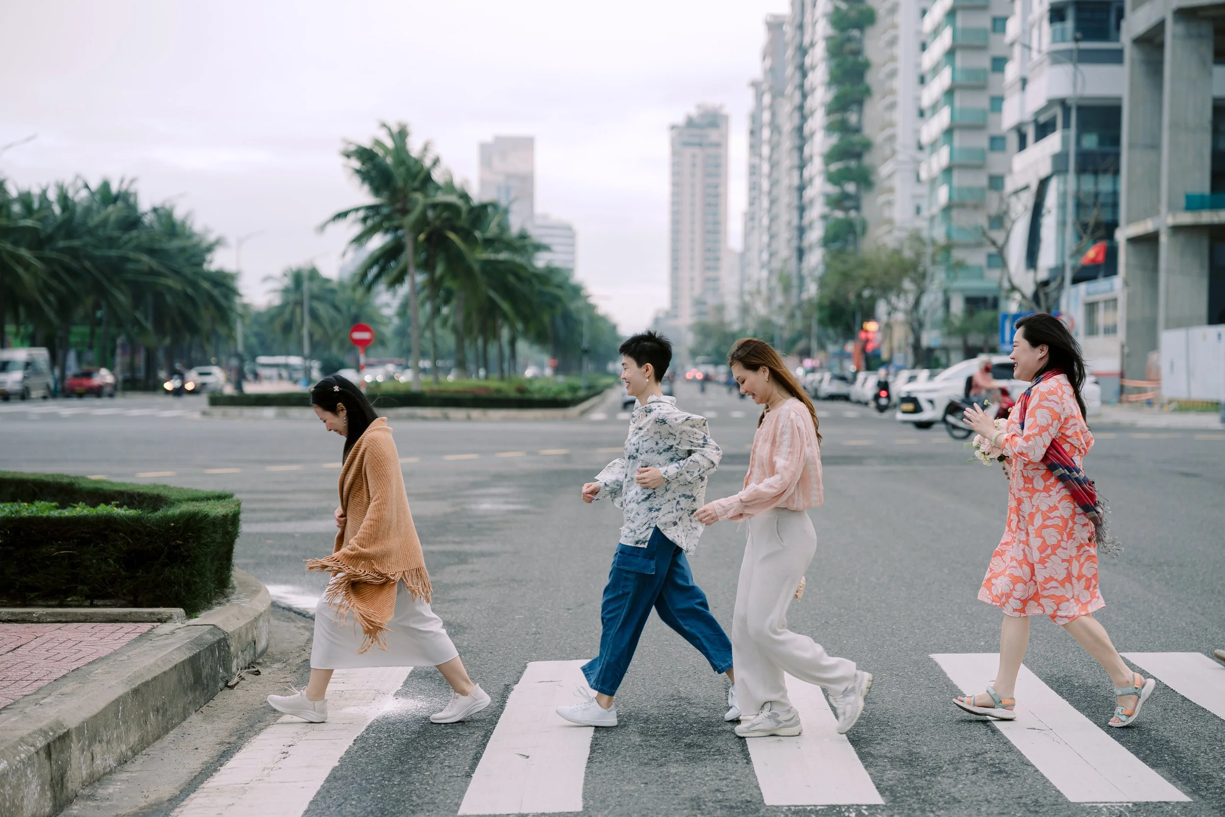 Four women walking across a city crosswalk on a cloudy day, with high-rise buildings and palm trees in the background.