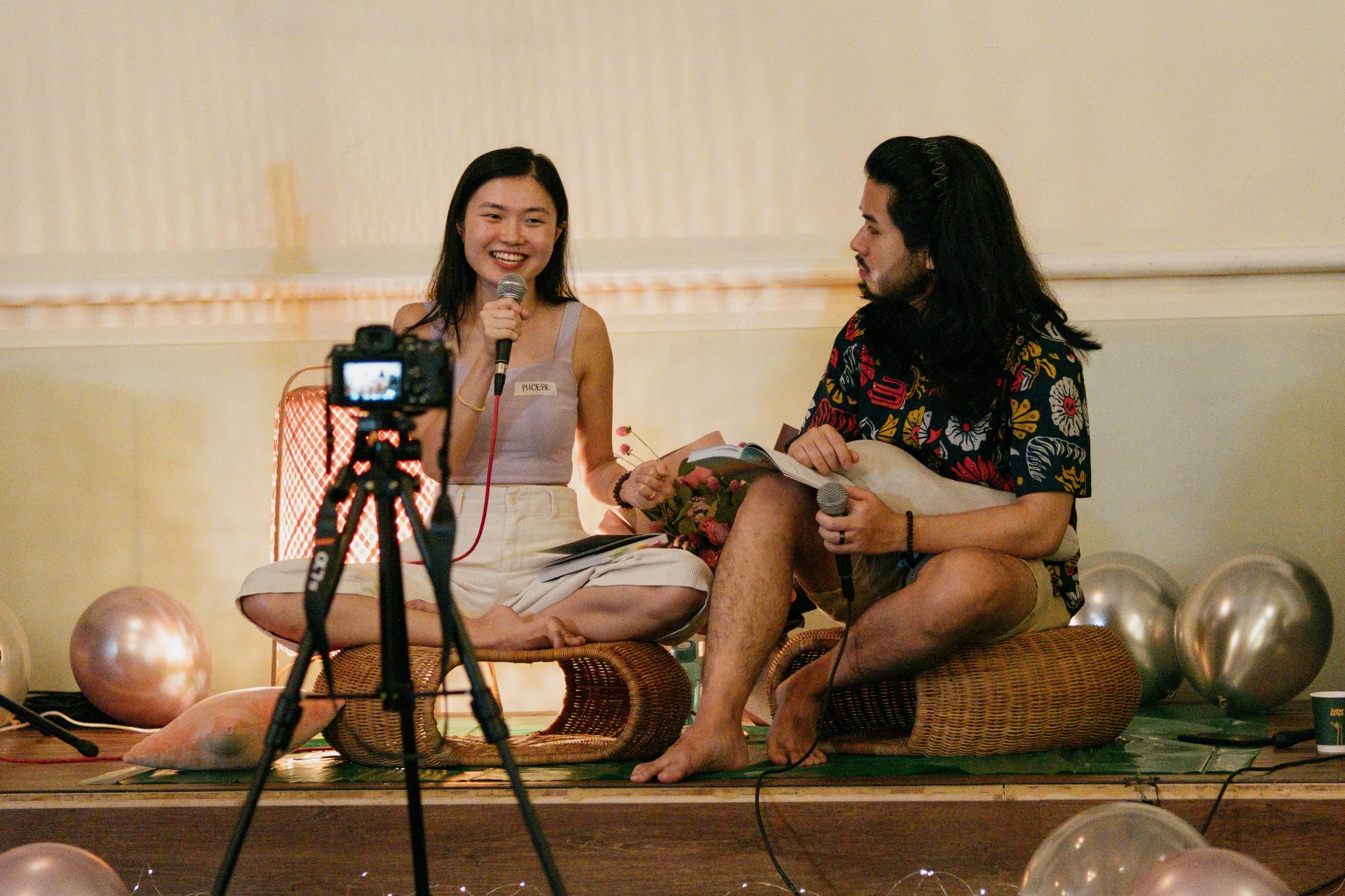 Two people sitting on floor cushions on a stage, one person holding a microphone and the other holding a book and a microphone, with balloons in the background.