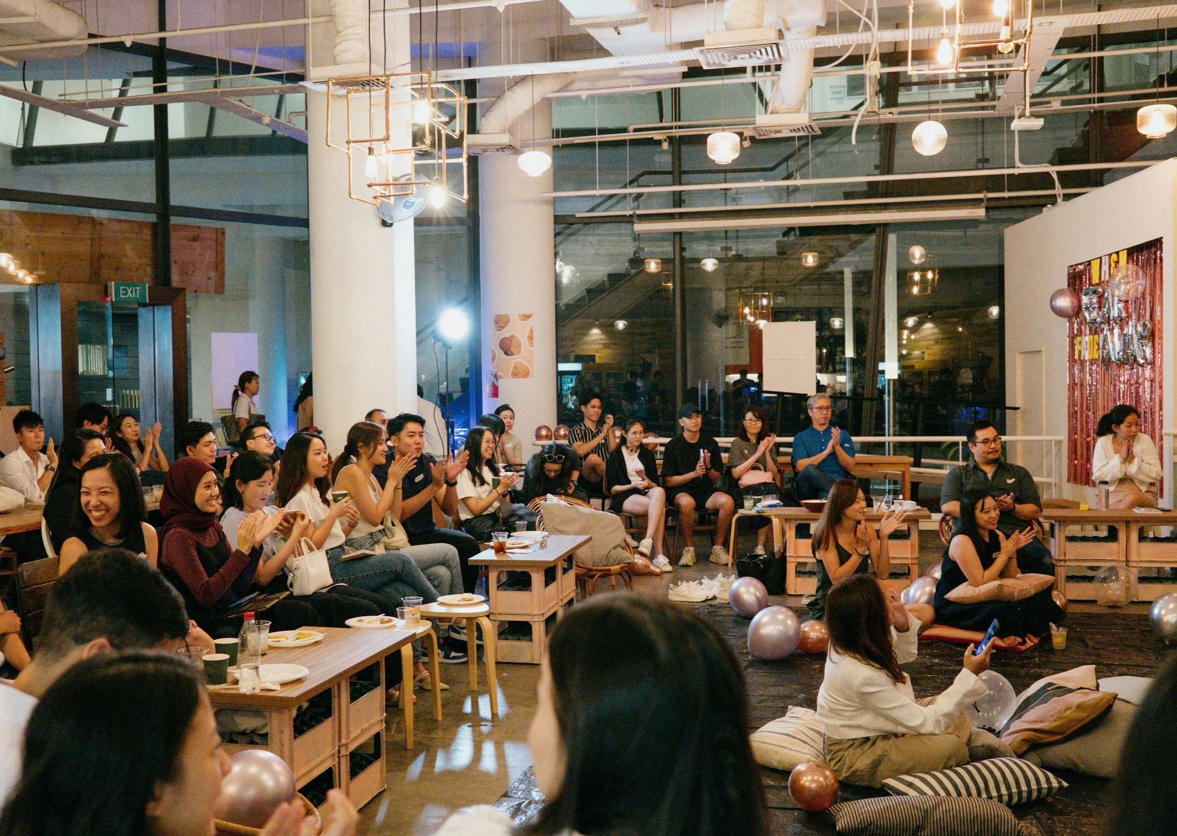 Group of people attending an indoor event or gathering, seated on benches and cushions, with some clapping and smiling, decorated with balloons and banners, in a modern, industrial-style venue with large windows and hanging lights.