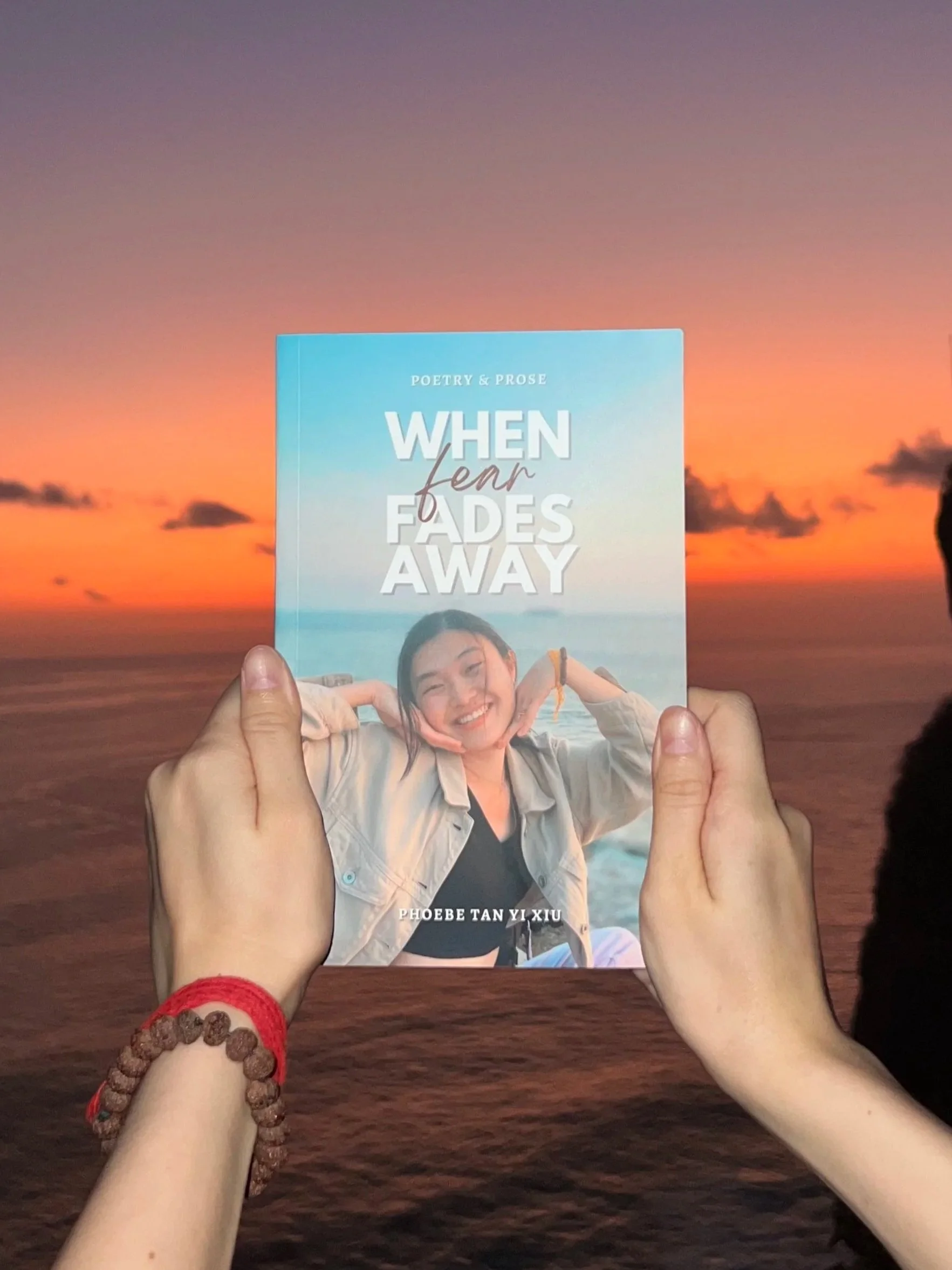 A person holding a book titled "When Fears Fade Away" by Phoebe Tan Yi Xiu in front of a sunset at the beach.