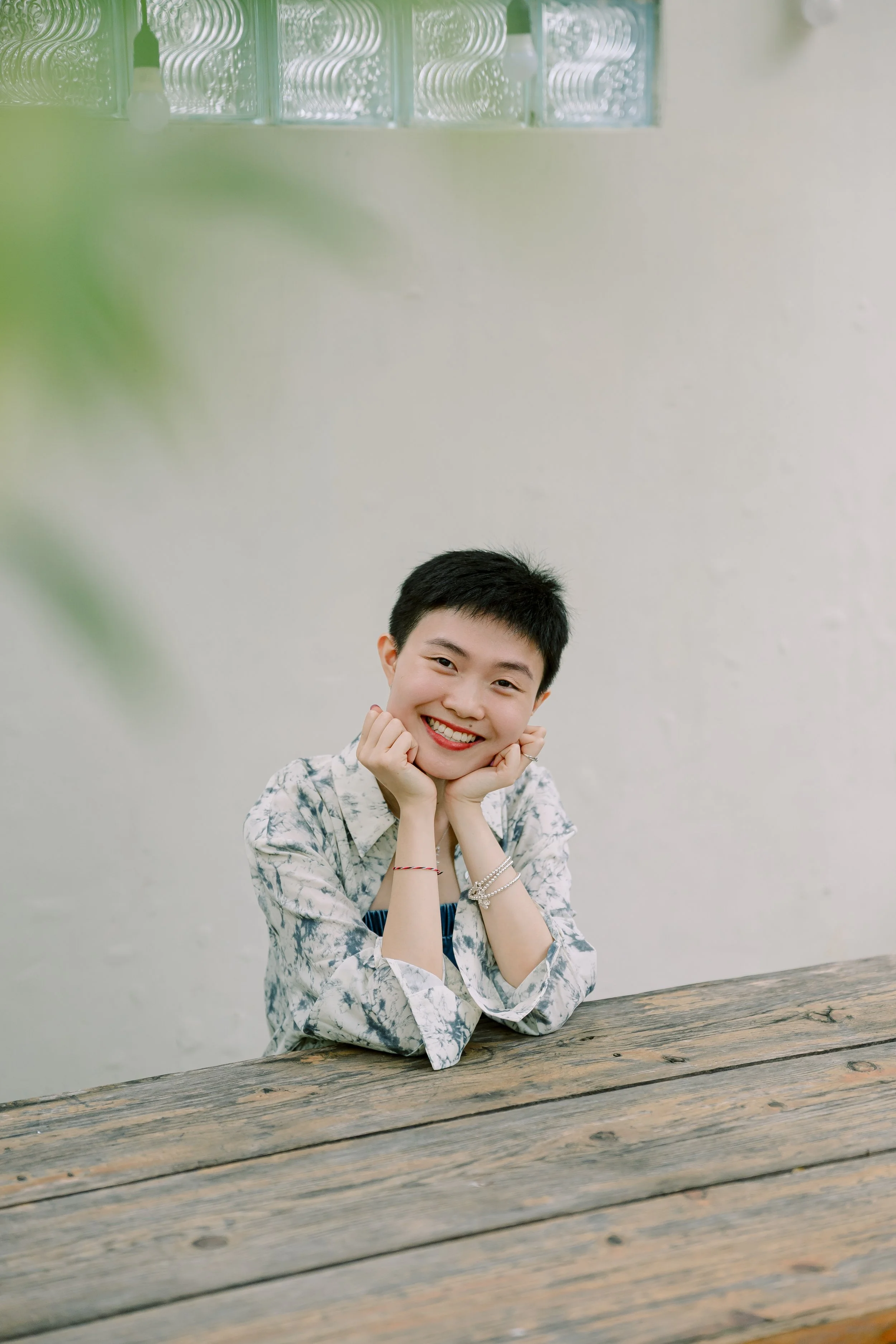 Smiling young woman with short black hair, wearing a patterned blouse with rolled-up sleeves, sitting at a rustic wooden table with a plain light-colored wall in the background.