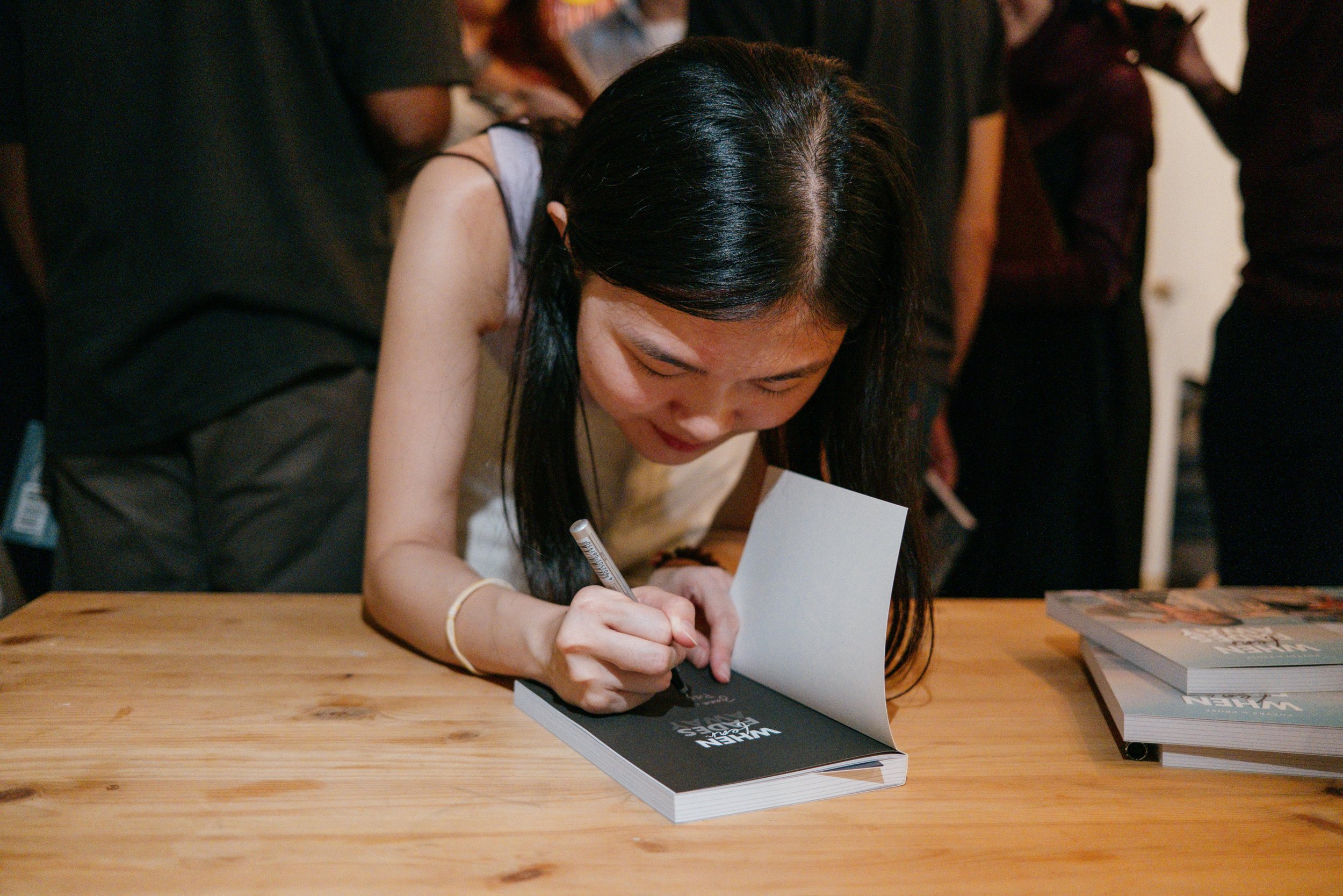Woman signing a book at a book signing event, with multiple people in the background.