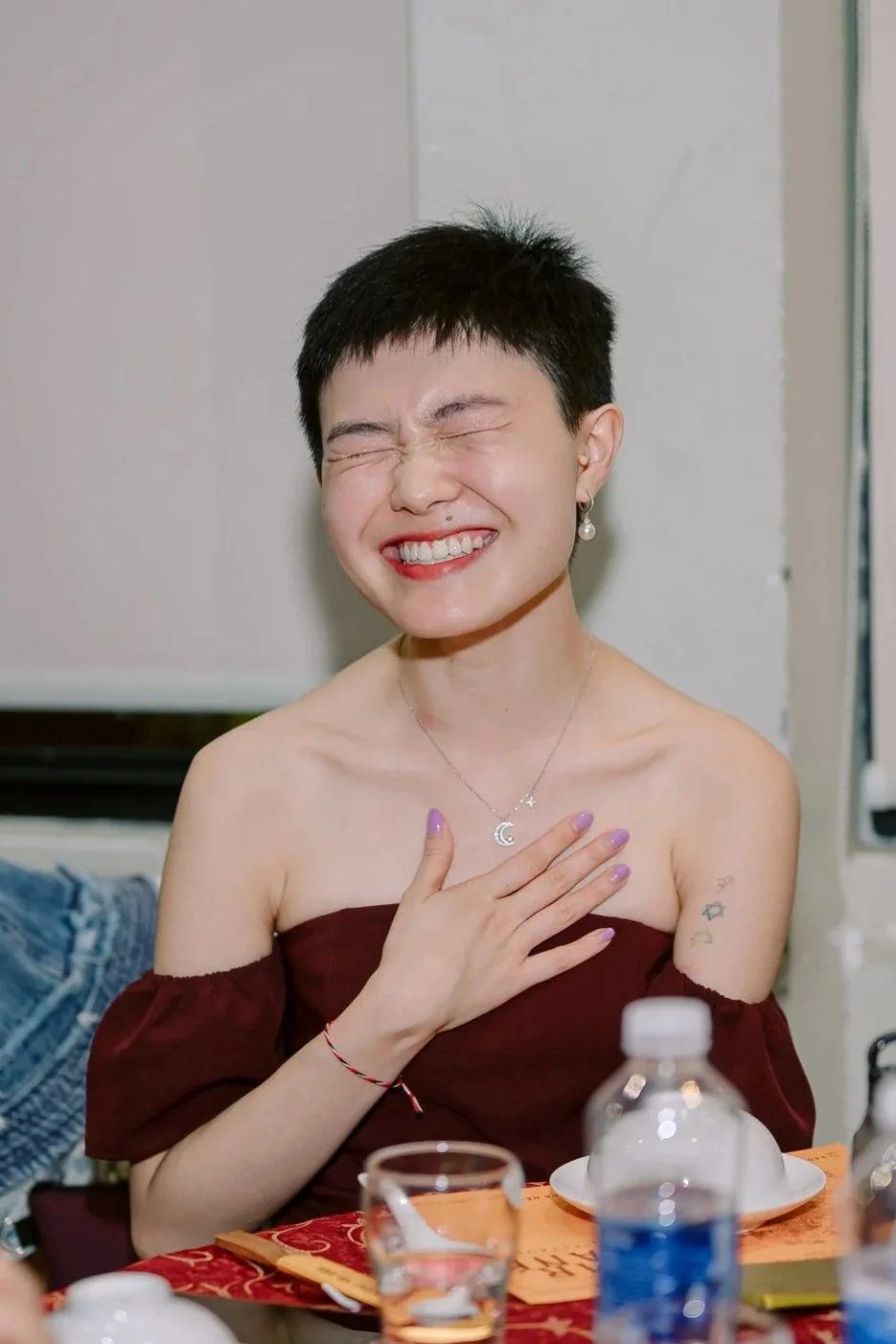 A young woman with short black hair, smiling widely with eyes closed, wearing a maroon off-shoulder top, silver moon and star necklace, pearl earrings, and purple nail polish, sitting at a table with plates, water bottle, and other items.
