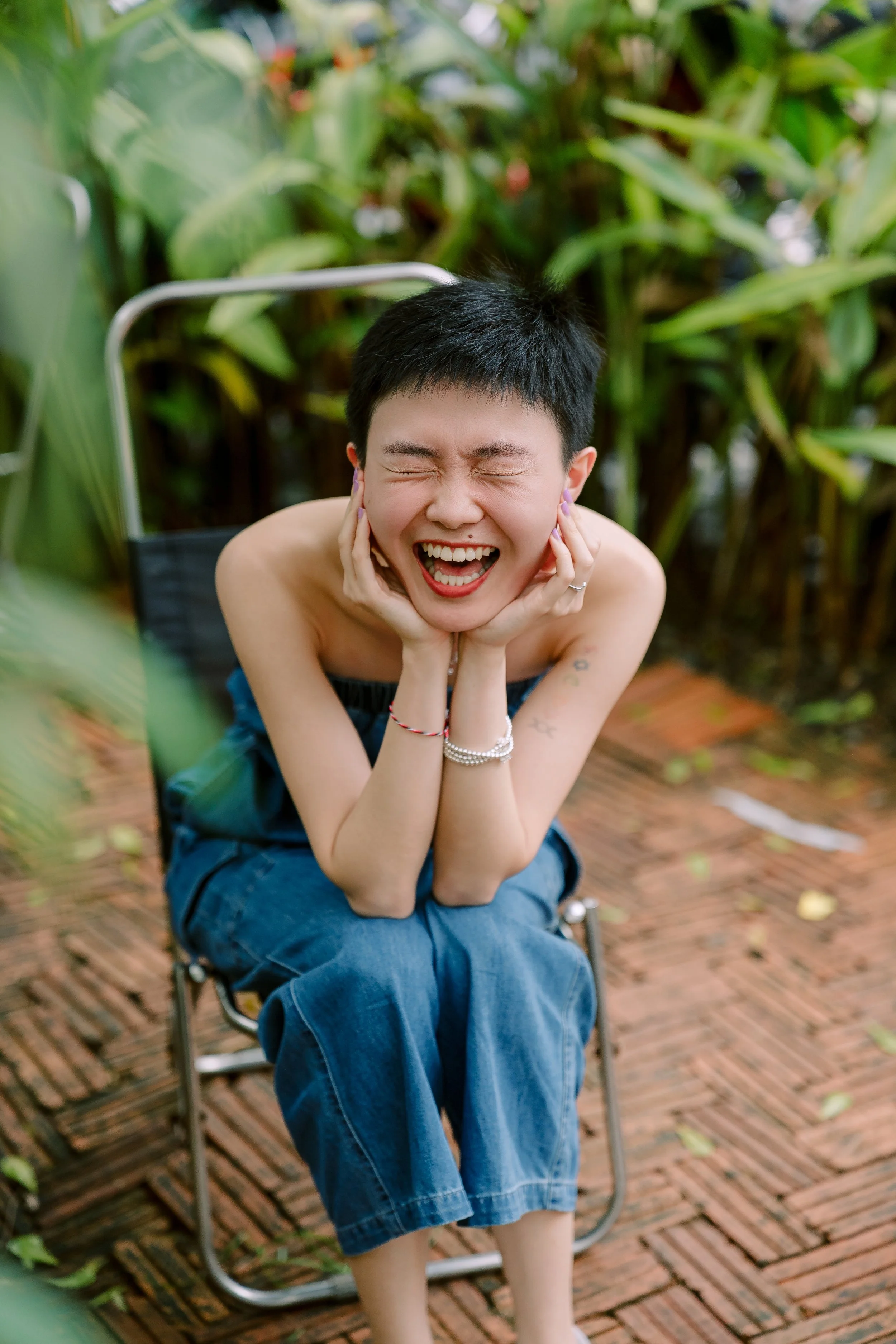 A woman with short hair laughing with her eyes closed, sitting outdoors on a brick pathway with green plants in the background.