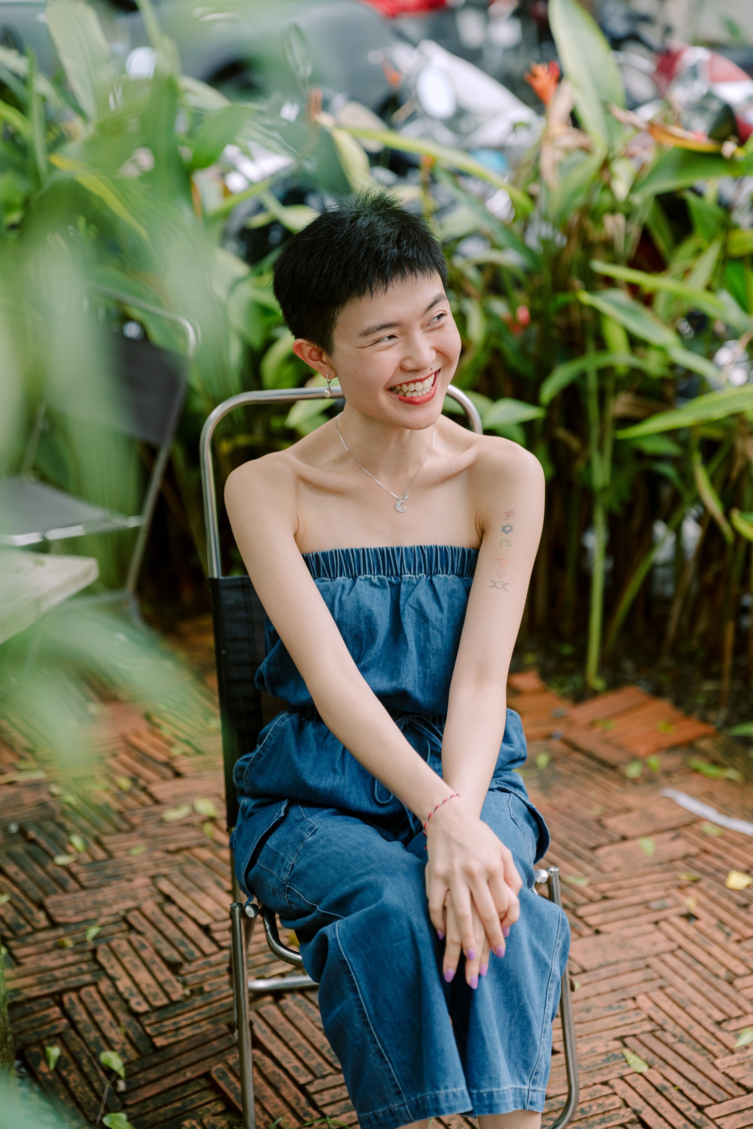 A smiling woman with short black hair and tattoos on her arm, wearing a strapless denim jumpsuit, sitting outside on a chair with green plants in the background.