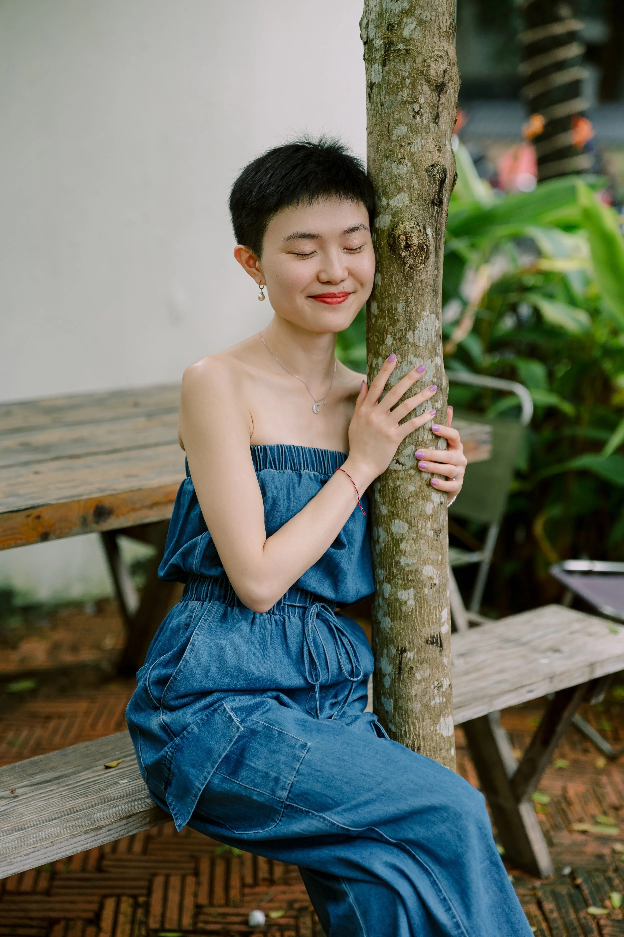 A young woman with short black hair, smiling with eyes closed, hugging a tree while sitting on a wooden bench outdoors, with green foliage in the background.