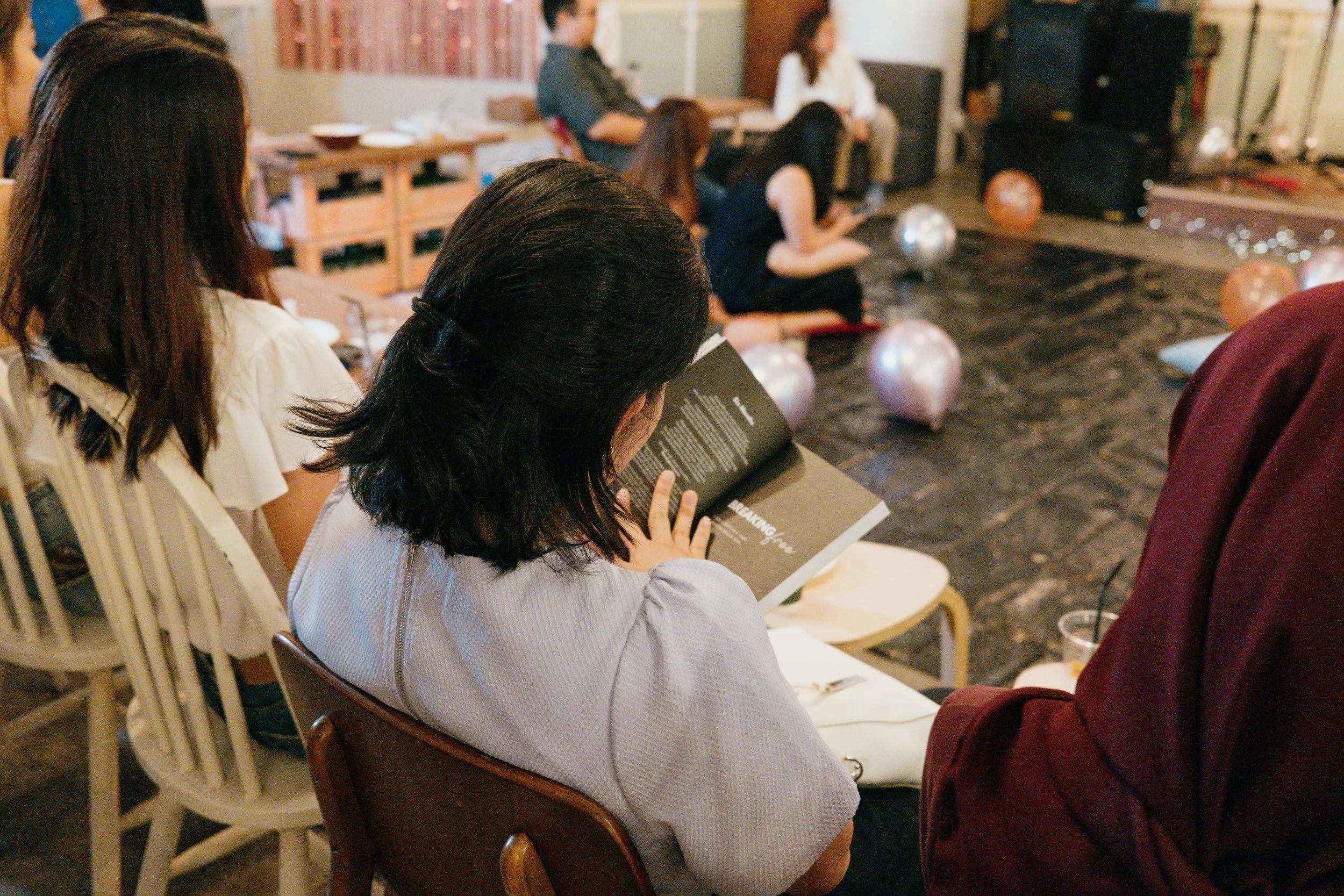 People seated in chairs, some reading, watching a karaoke performance with balloons on the stage.