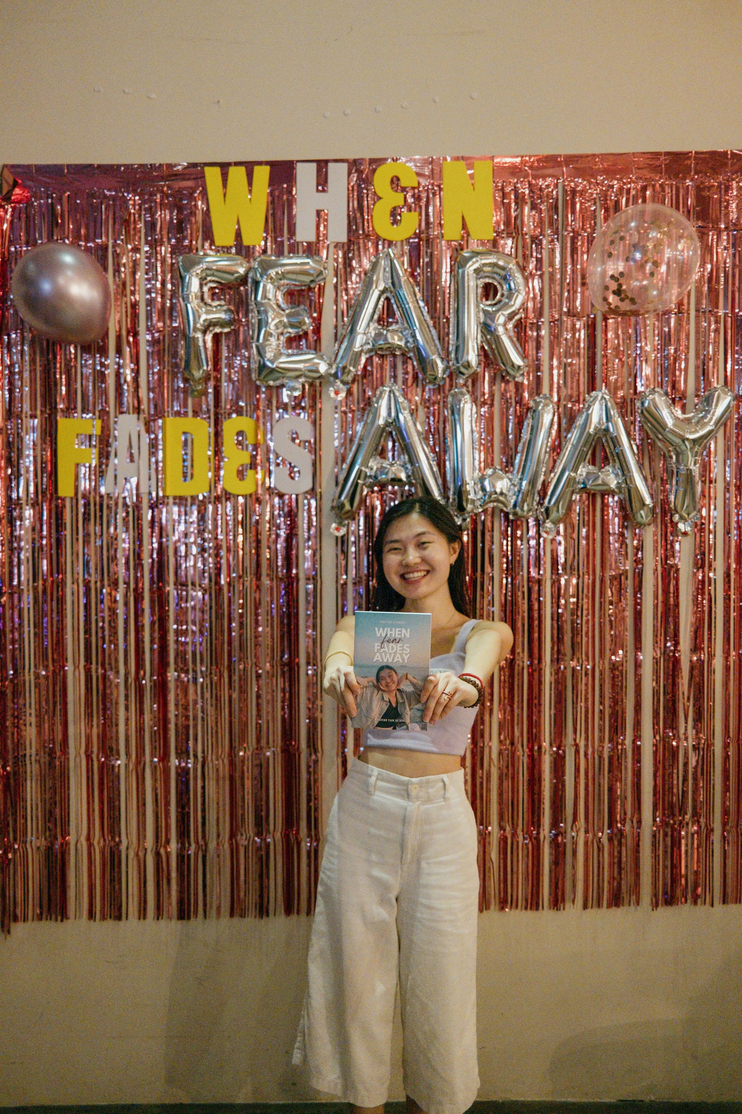 Young woman standing in front of a decorative backdrop with balloons and shiny banner, holding a book titled 'When Fades Away' and smiling.