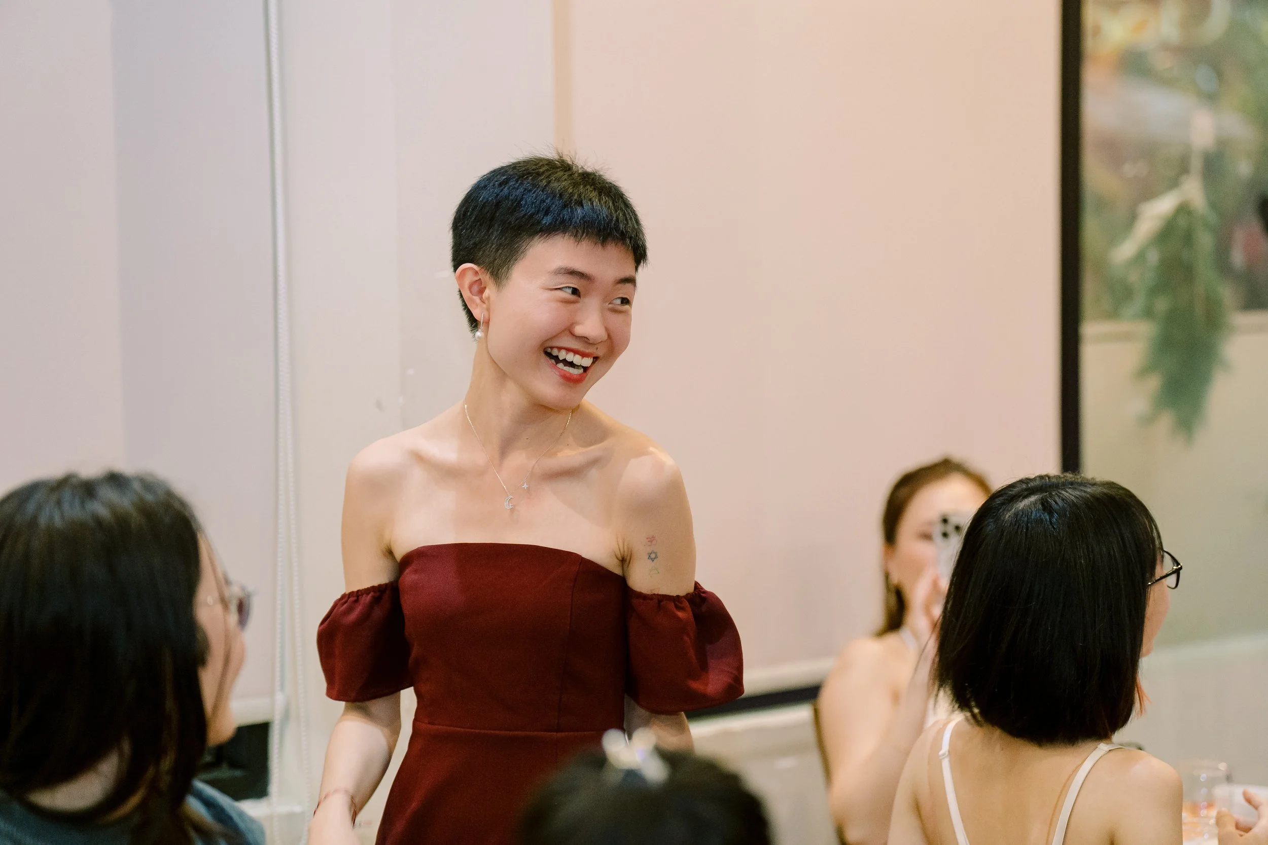 A woman with short black hair, wearing a strapless maroon dress, smiling while talking to others in a social gathering.
