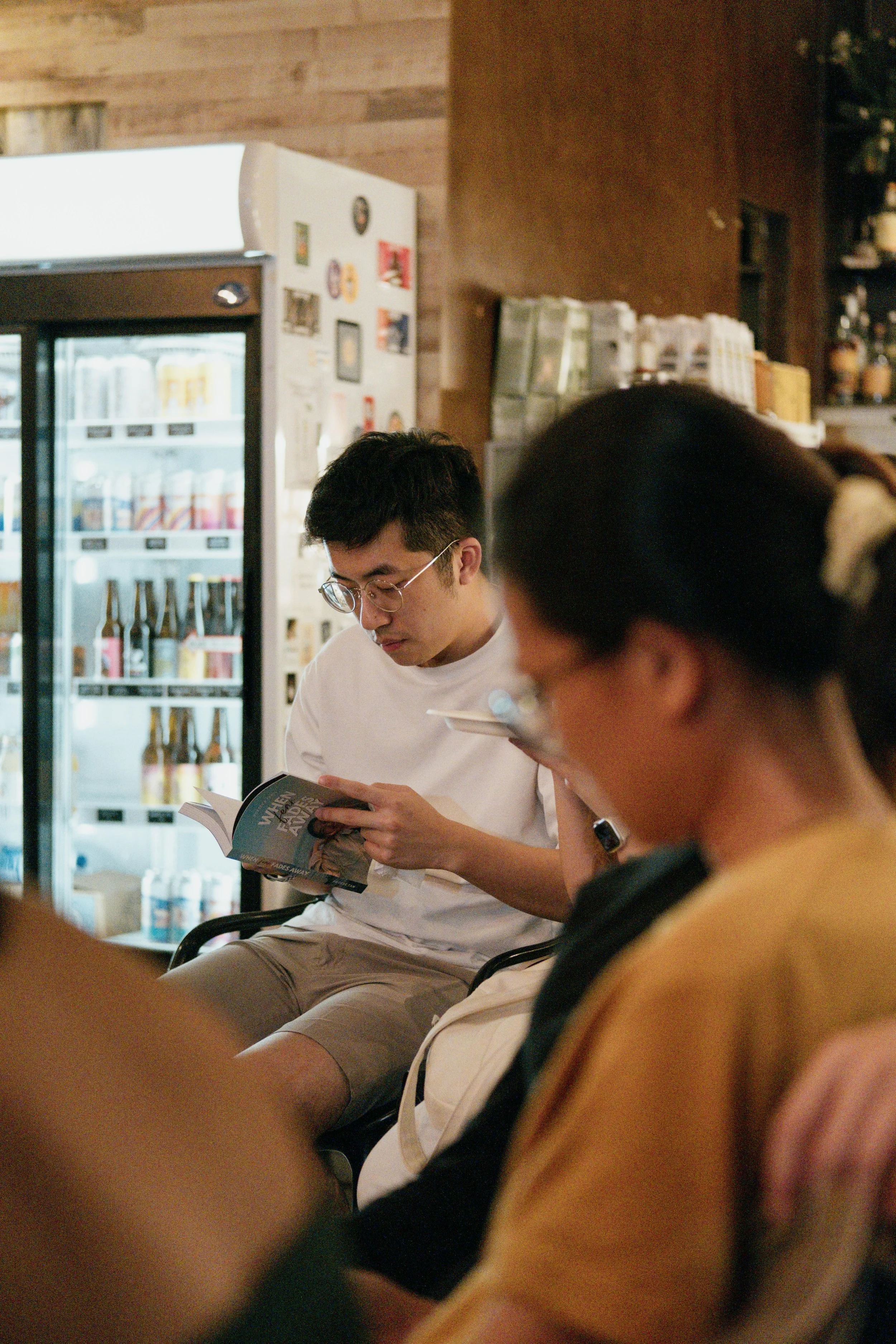 A young man with glasses reading a book while sitting in a cafe, with a fridge of drinks behind him and a wooden wall.