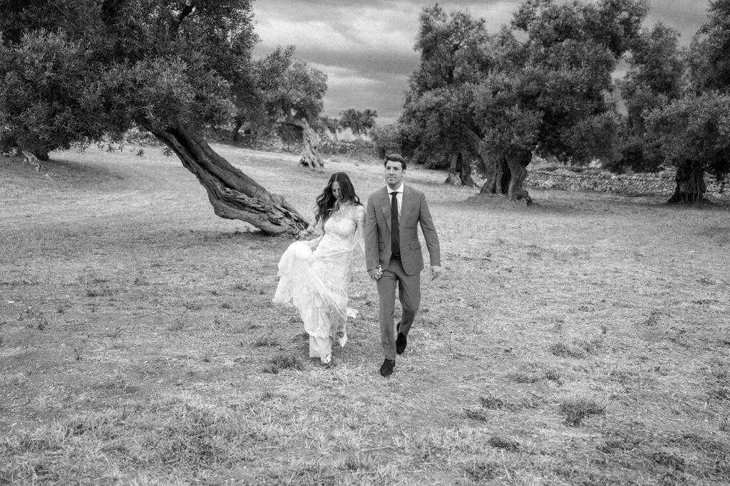 A bride and groom walking hand in hand through a landscape with trees, captured in black and white.