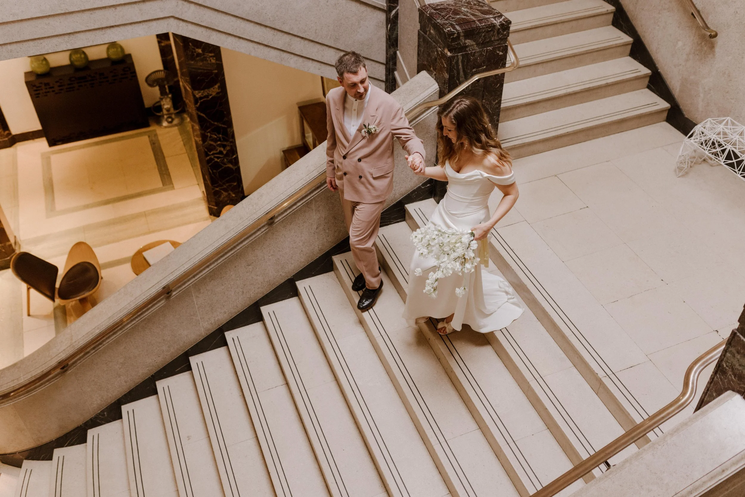 A bride and groom holding hands on a staircase in a hotel or building, with the bride wearing a white wedding dress and holding a bouquet of white flowers.