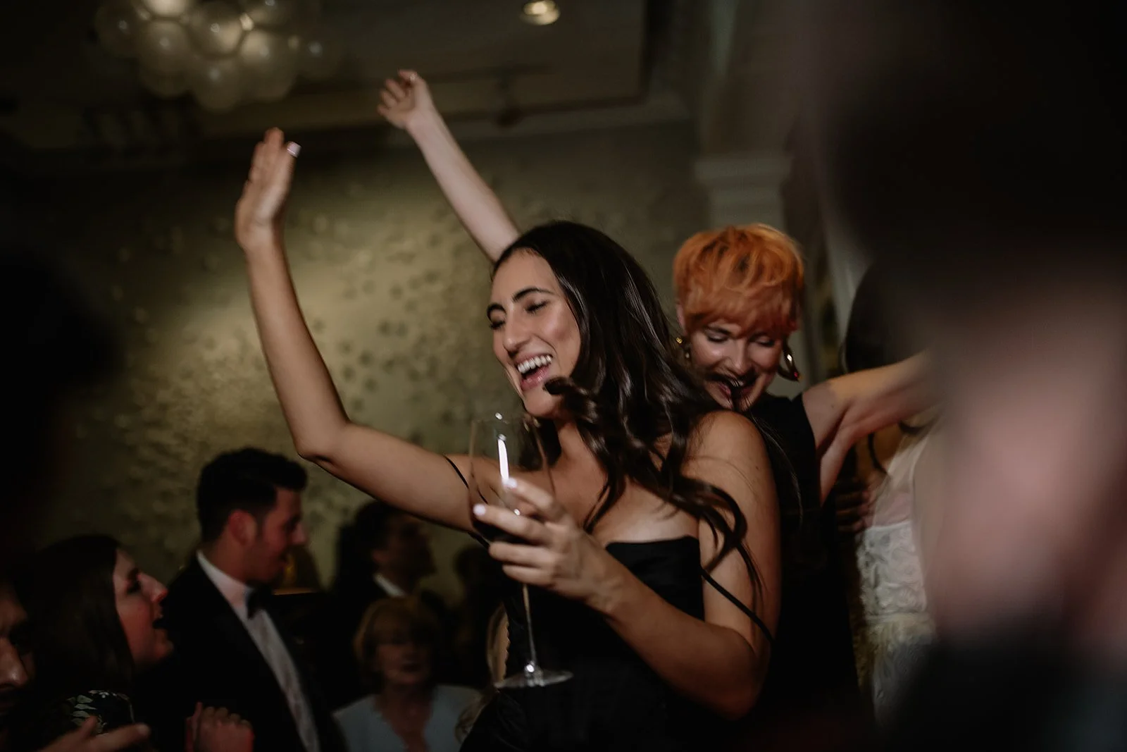 Women dancing and smiling at a celebration, with other guests dressed formally in the background.