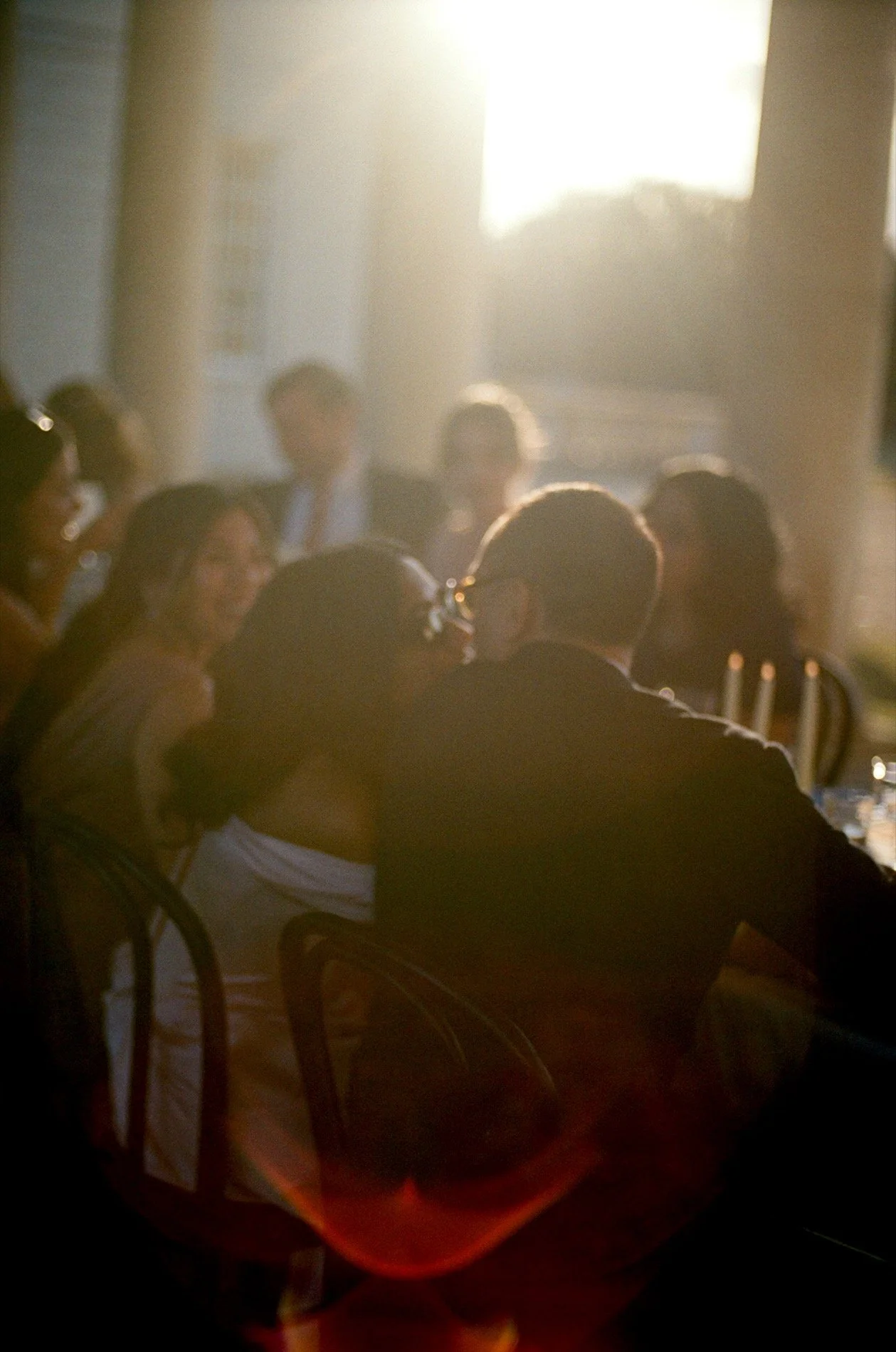 People sitting at a formal gathering or banquet, illuminated by soft, warm lighting with sunlight streaming through a window behind them.