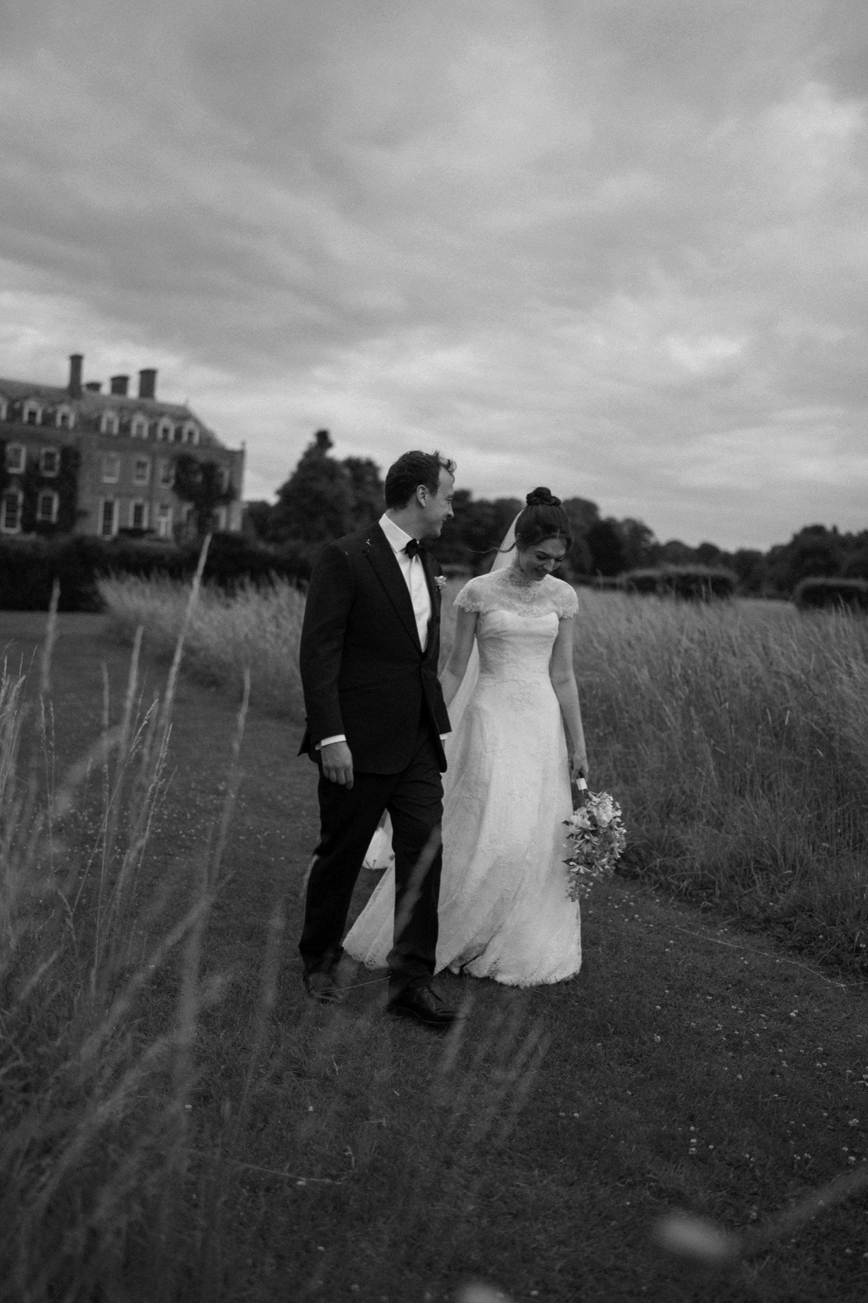 Black and white photo of a bride and groom walking together outdoors, with the bride holding a bouquet, during an overcast day near a large house or estate.