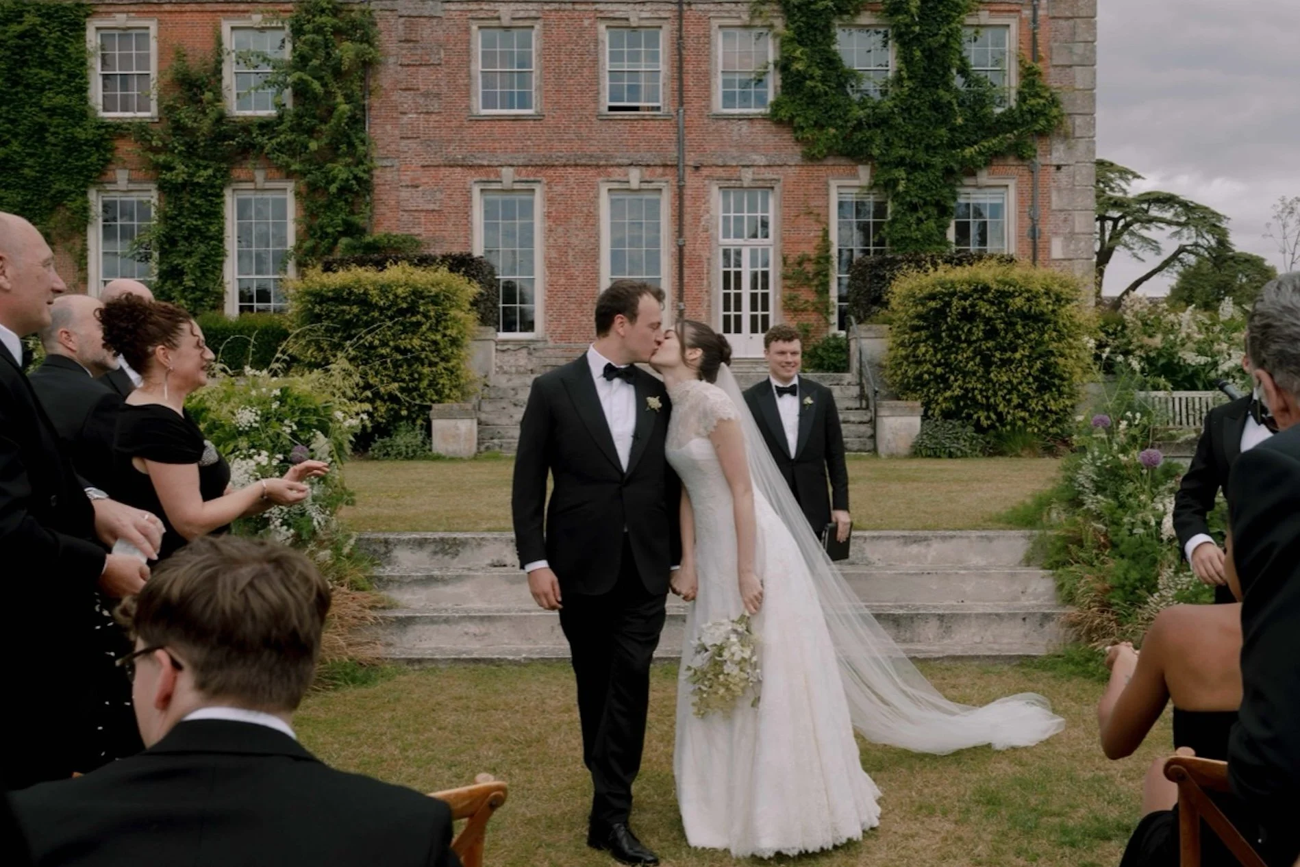 A bride and groom kiss at their outdoor wedding ceremony in front of a brick building. The bride wears a white wedding dress with a long veil, holding a bouquet. The groom is dressed in a black tuxedo. Friends and family in formal attire observe; some are in black suits and dresses. The scene includes greenery, flowers, and steps leading to the building.