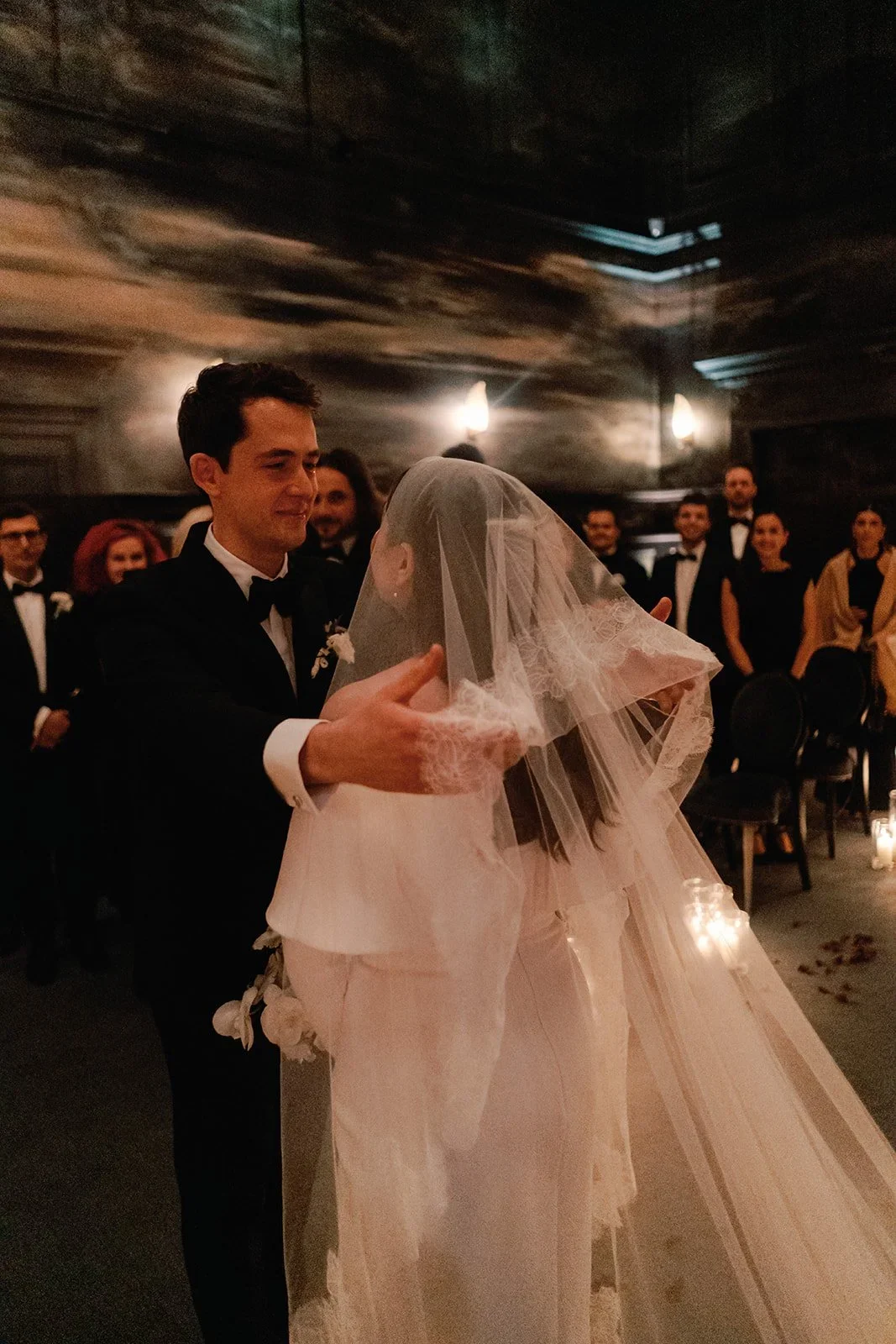 A bride and groom sharing a dance at their wedding reception. The groom is dressed in a black tuxedo and the bride wears a long, flowing veil and gown. Guests in formal attire are watching them, and the venue has dark walls with soft lighting.