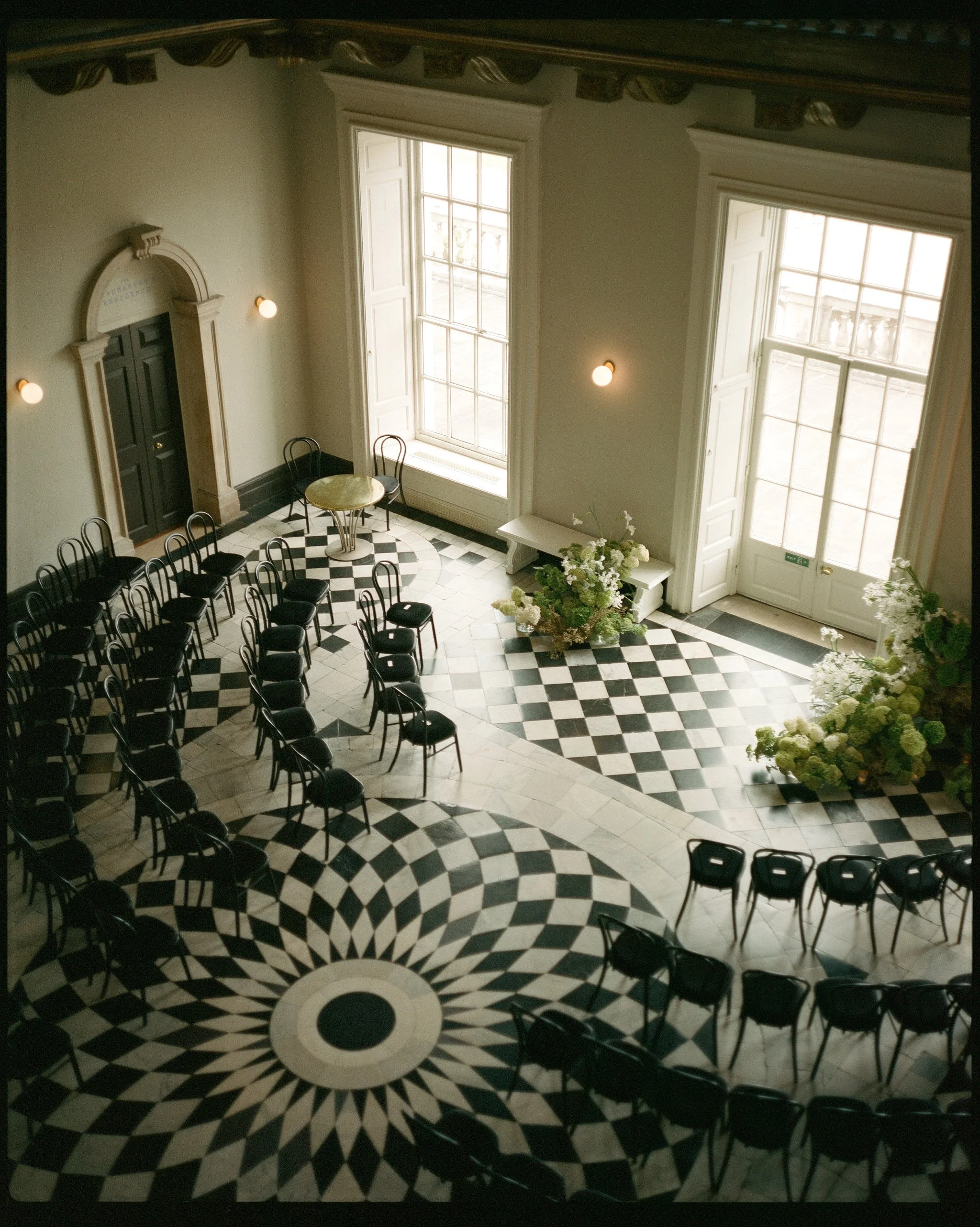 An elegant, spacious room with black and white checkered marble floors, large tall windows letting in natural light, and black chairs arranged in rows, suggesting it is set up for a ceremony or event. There are also floral arrangements of white and green flowers near the entrance.