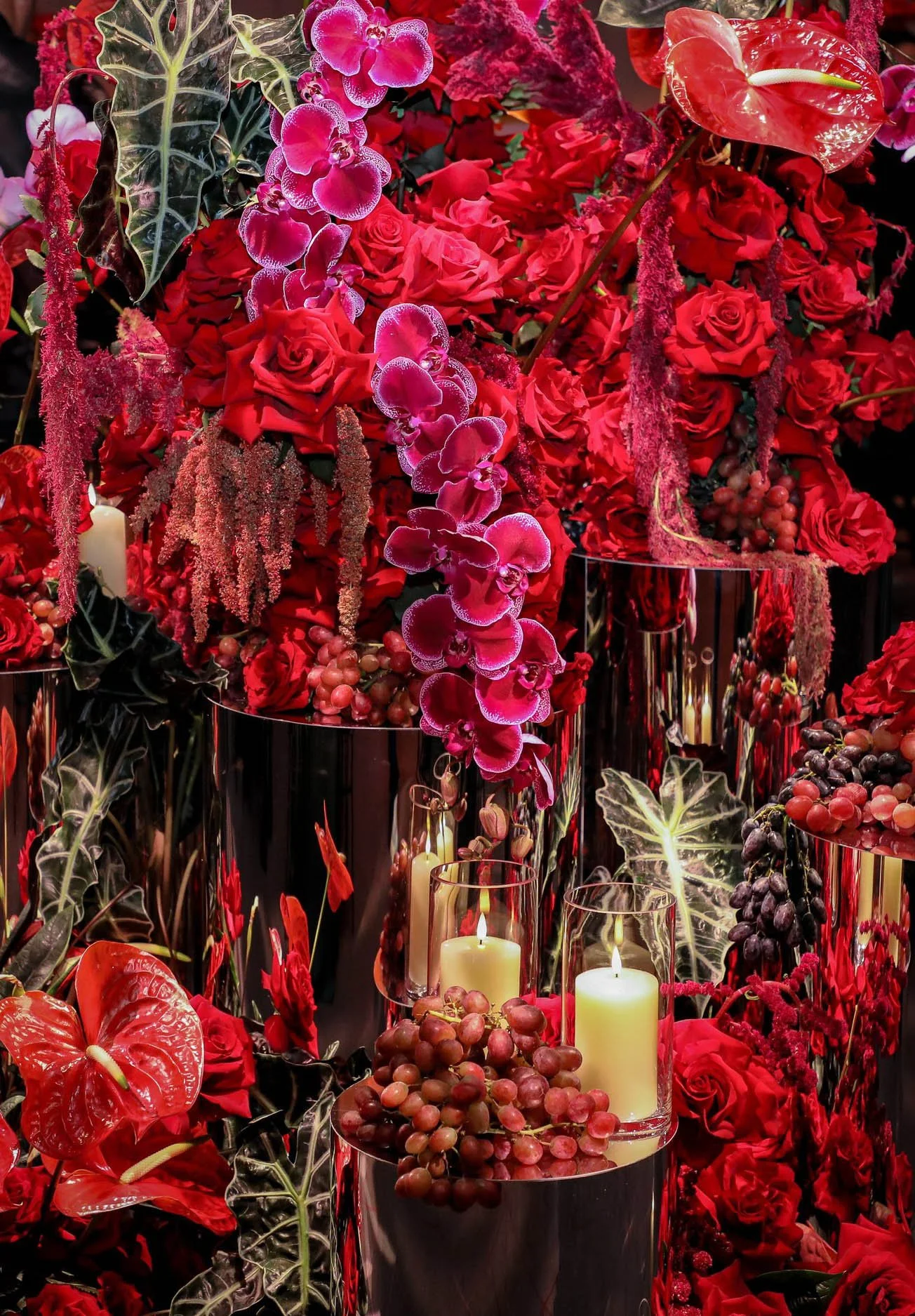 A floral arrangement with red roses, purple orchids, red anthuriums, green leafy plants, and candles in glass holders.