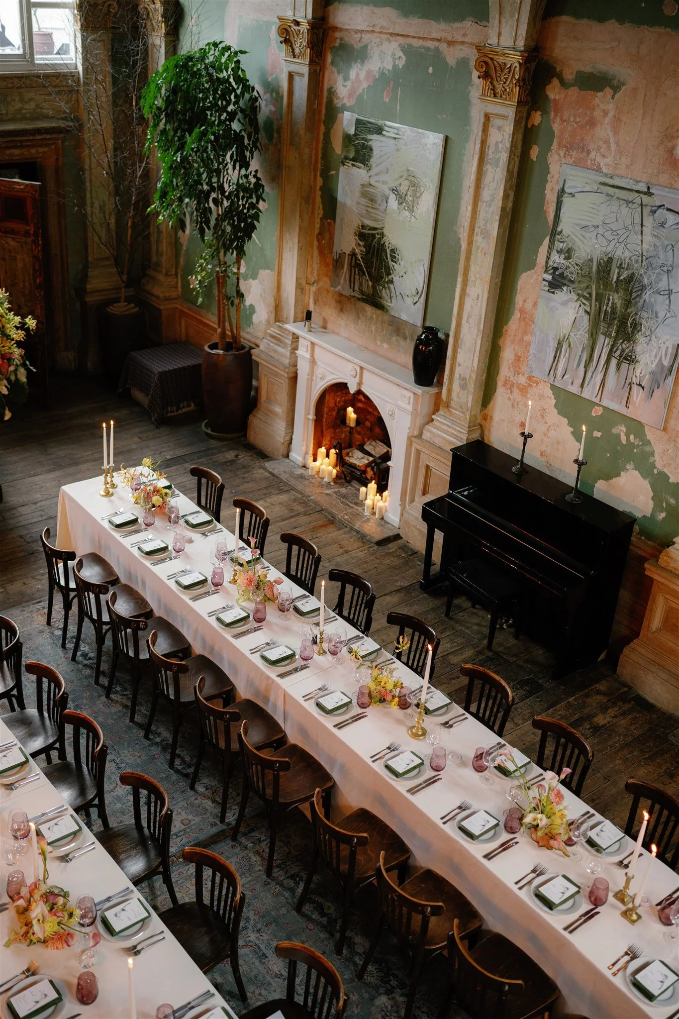 Long dining table set with plates, pink goblets, silverware, and floral centerpieces, in a decorated room with paintings, tall candles, a fireplace with candles, a black piano, and large potted plant.