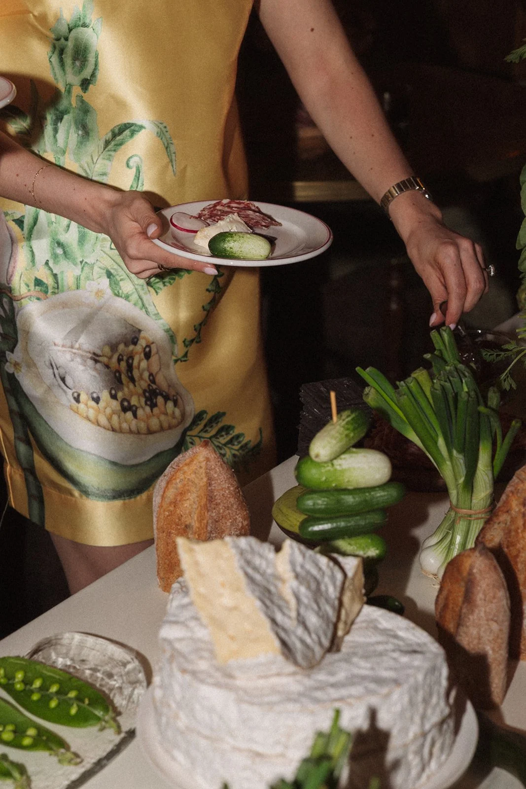 Close-up of a table with cheese, bread, green onions, and cucumbers, with a person holding a plate with cured meat, cheese, and a radish in the background.
