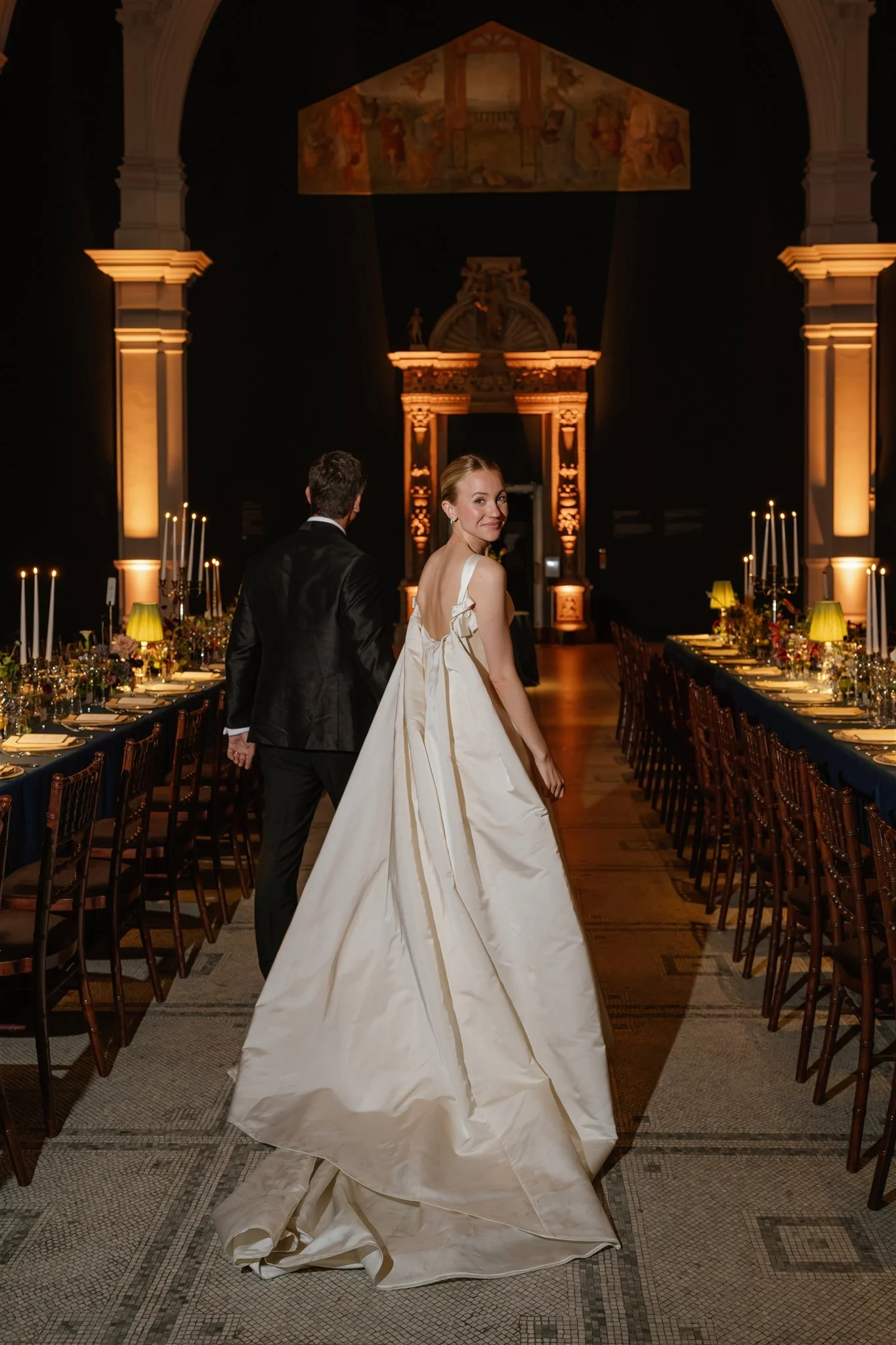 A bride in a white wedding gown and a groom in a black tuxedo standing in a beautifully decorated reception hall with long tables, candles, and elegant lighting.
