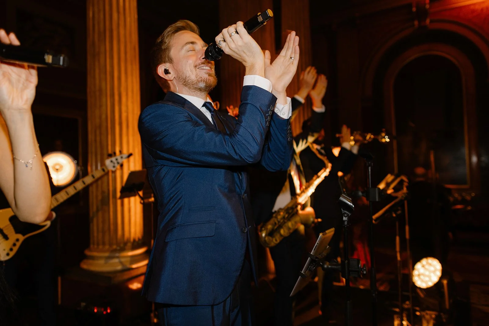 A man in a blue suit singing into a microphone during a live music performance with band members playing instruments in the background, in a dimly lit venue.