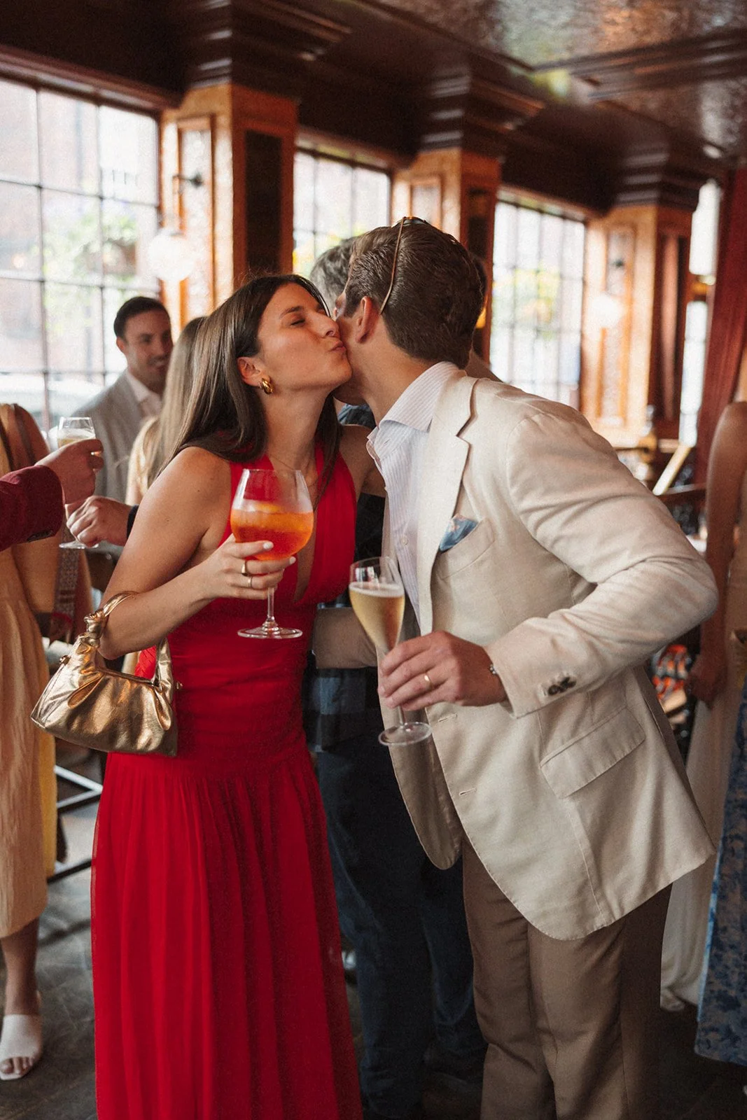 A woman in a red dress and gold handbag kisses a man in a beige suit, both holding drinks, at a social gathering.