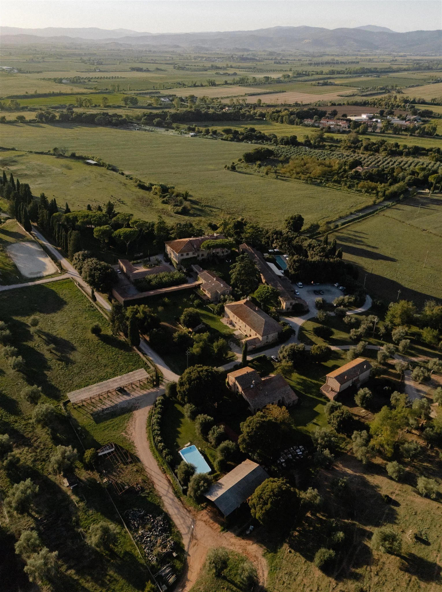 Aerial view of a rural estate with multiple buildings, swimming pool, gardens, trees, and surrounding farmland and fields extending to distant mountains.