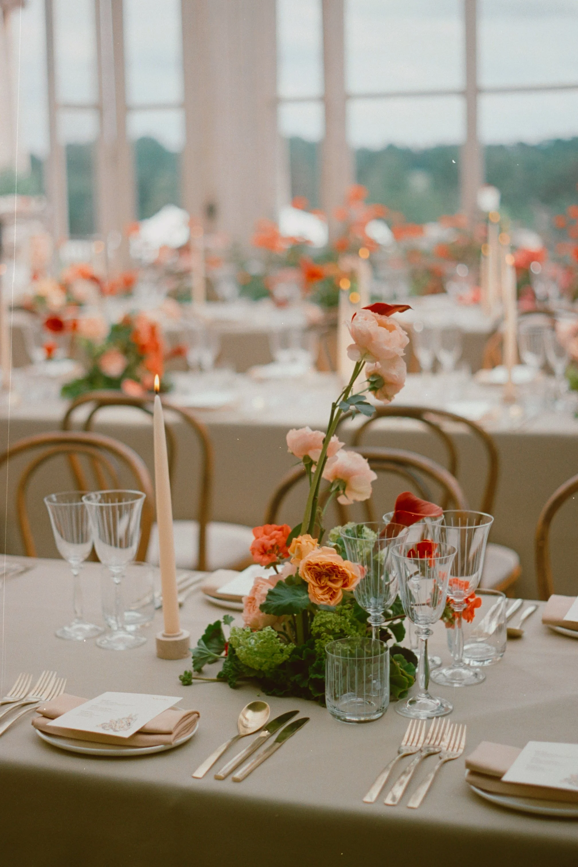 Elegant wedding table setting with pink and orange flowers centerpiece, candle, glassware, cutlery, and menu cards on a beige tablecloth, with a window and outdoor scenery in the background.