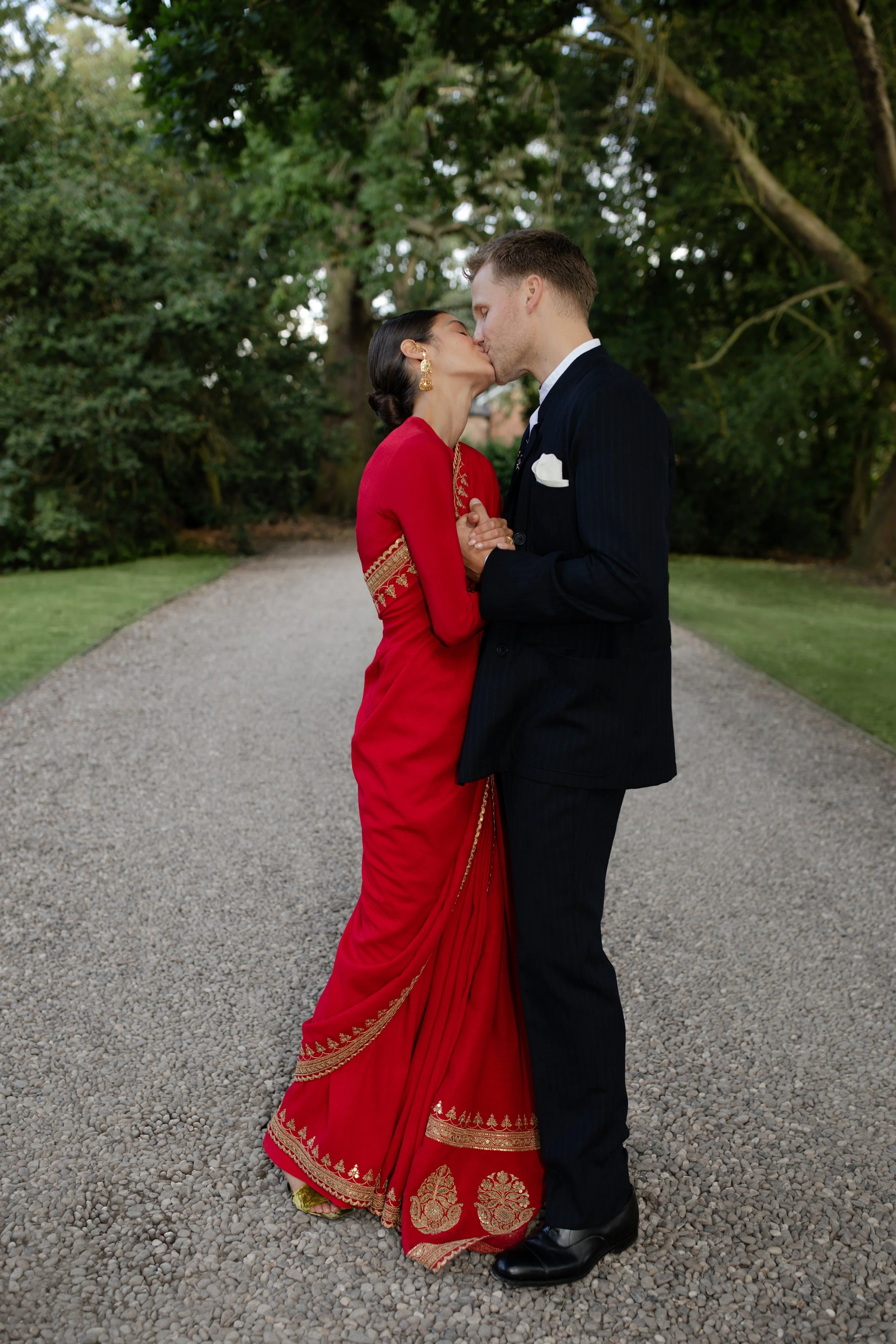 A couple in formal attire sharing a kiss on a gravel path surrounded by trees.