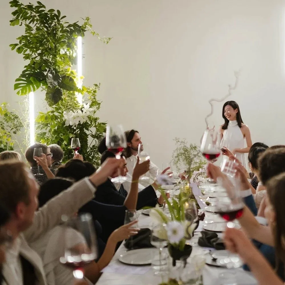 People at a celebration dinner raising glasses of red wine, with a woman standing and smiling near the head of the table, likely giving a toast, in a bright, modern room decorated with plants.