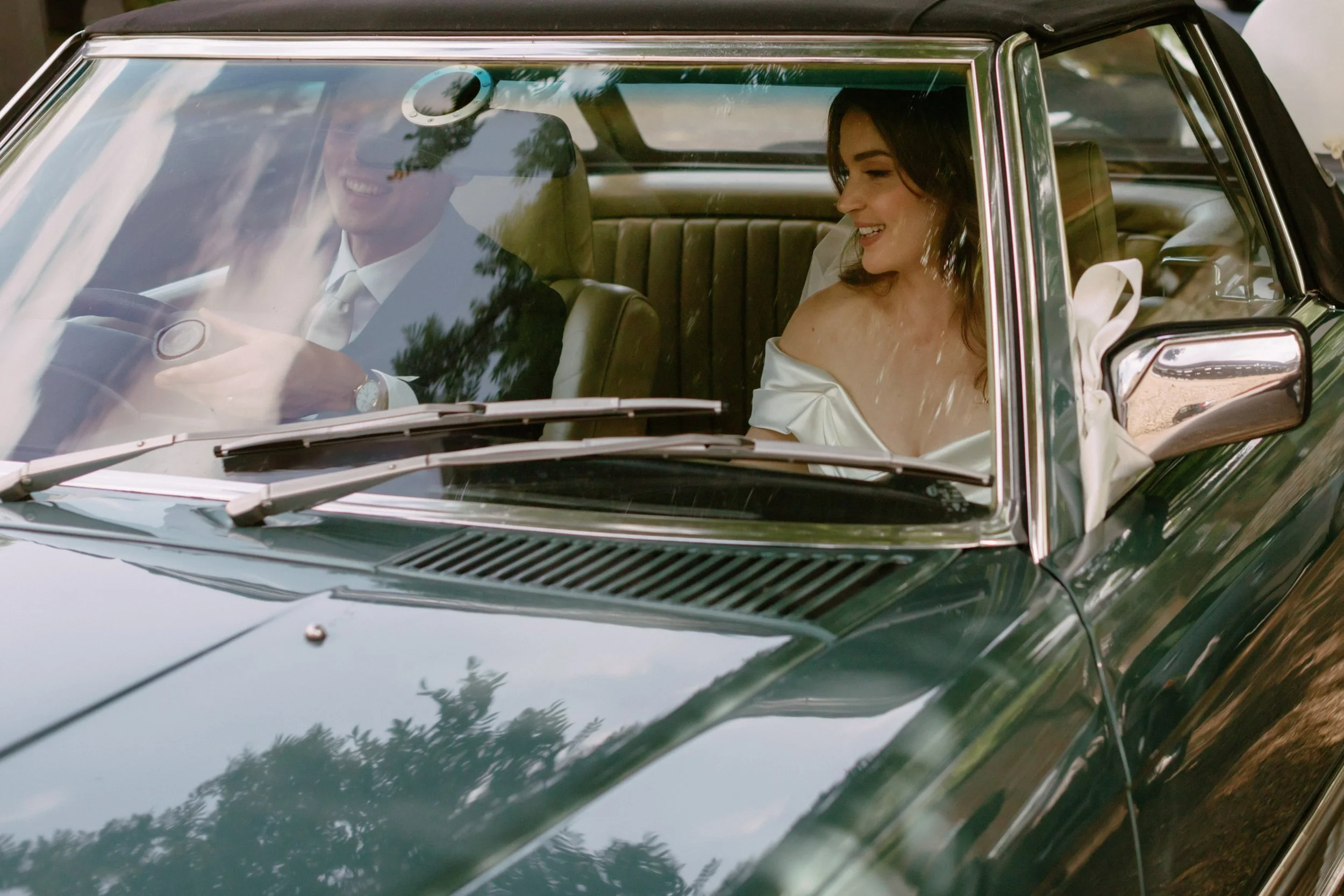 A bride and groom sitting in a vintage green car, smiling and talking to each other. The bride is wearing an off-shoulder wedding dress and the groom is in a suit, with trees and reflections visible on the car's windshield.