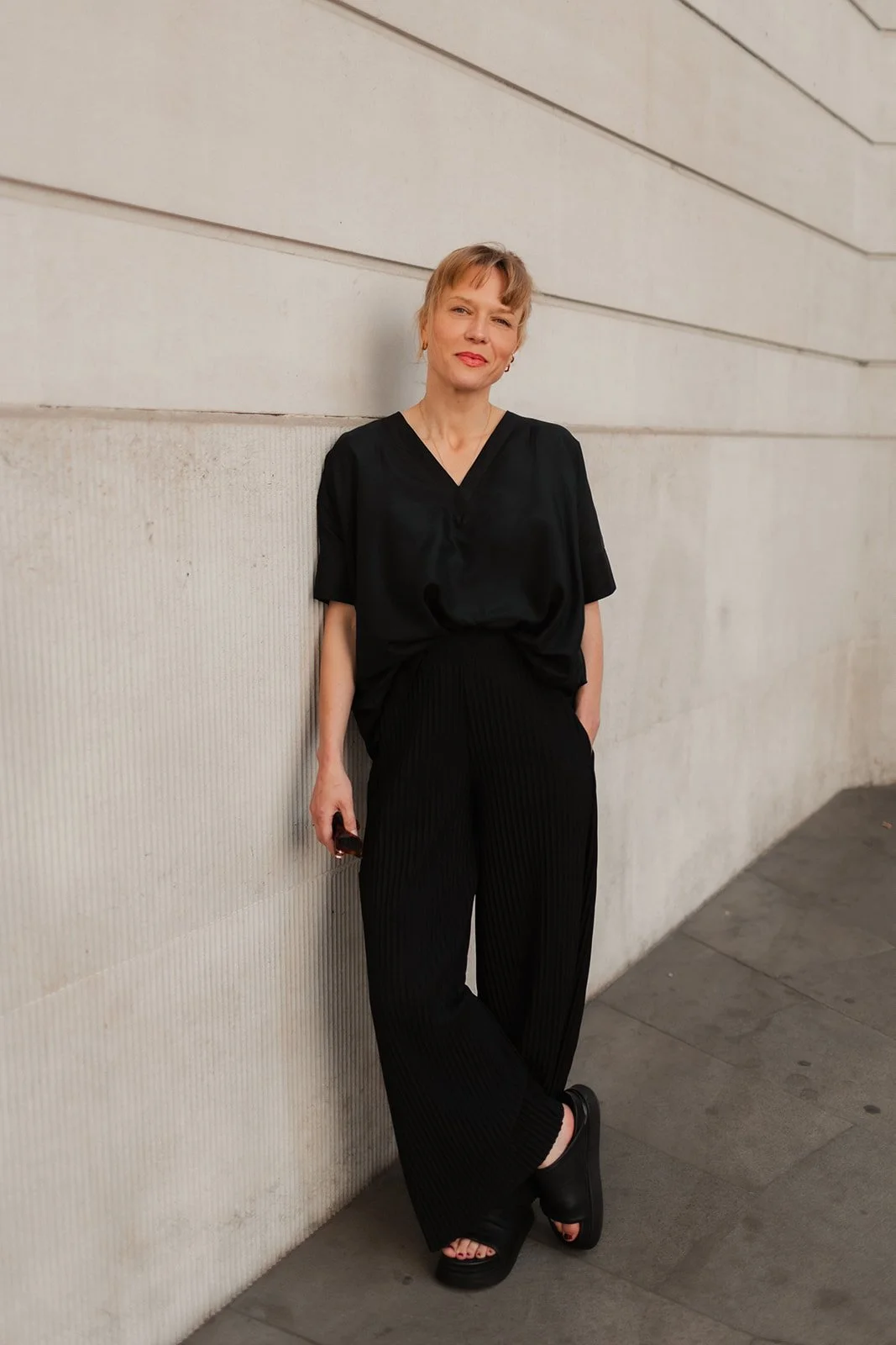 A woman in black outfit standing against a light-colored building wall, looking at the camera with a slight smile.
