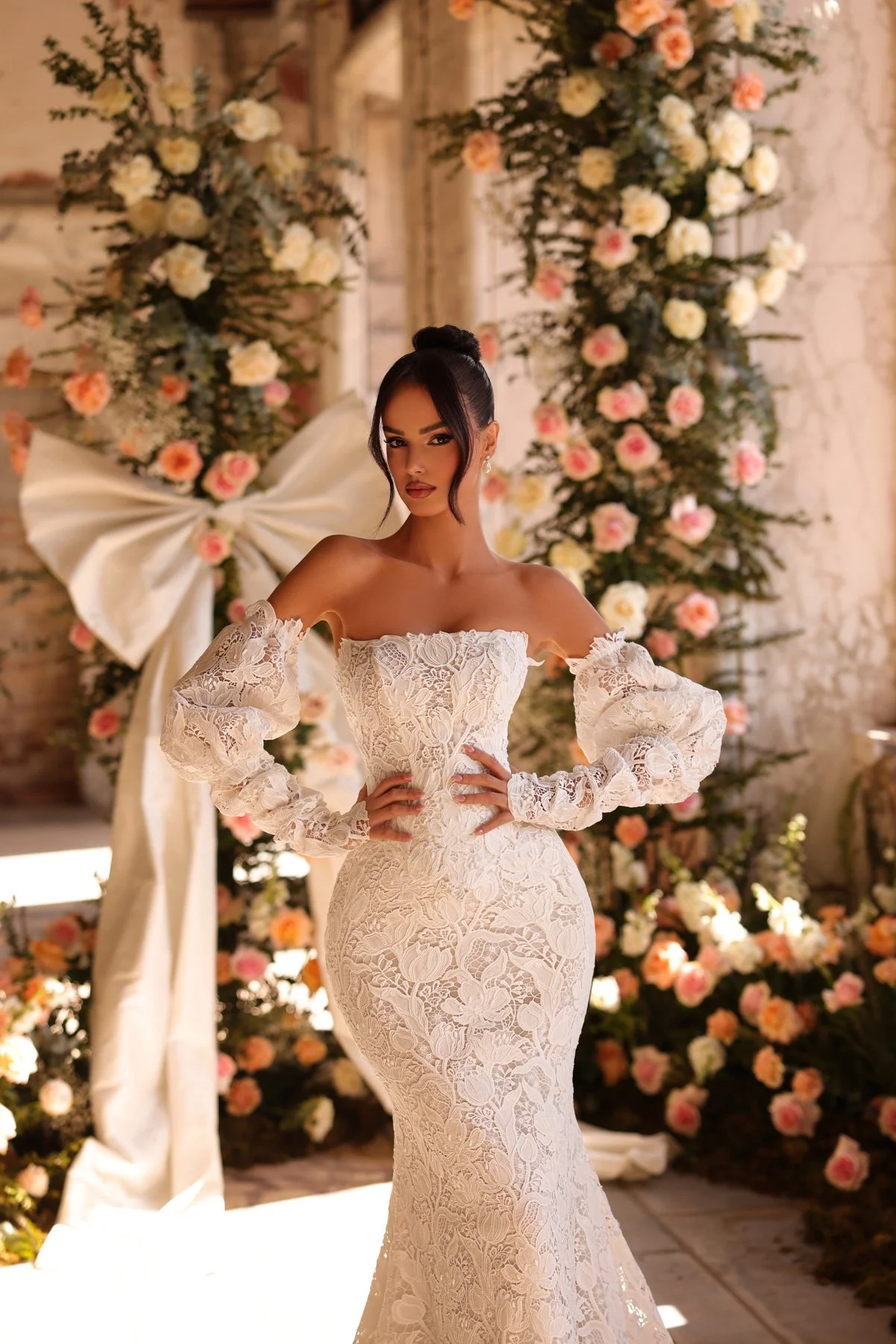 Mulher em vestido de noiva de renda, com cabelo preso em coque, posando diante de uma decoração floral.